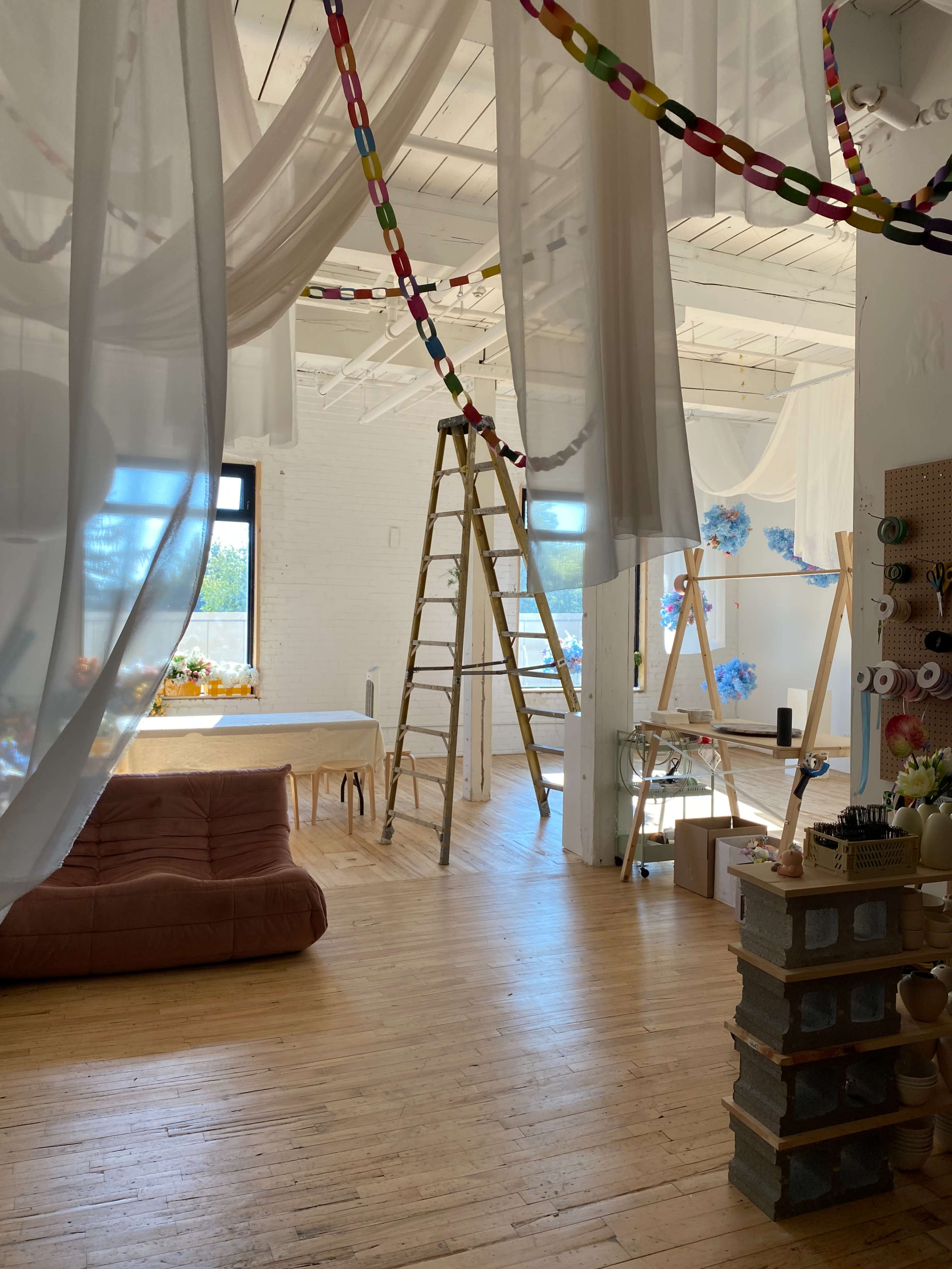 The image shows a spacious room with wooden floors, draped fabric, a ladder, and various decorations, including colorful chains and floral arrangements.