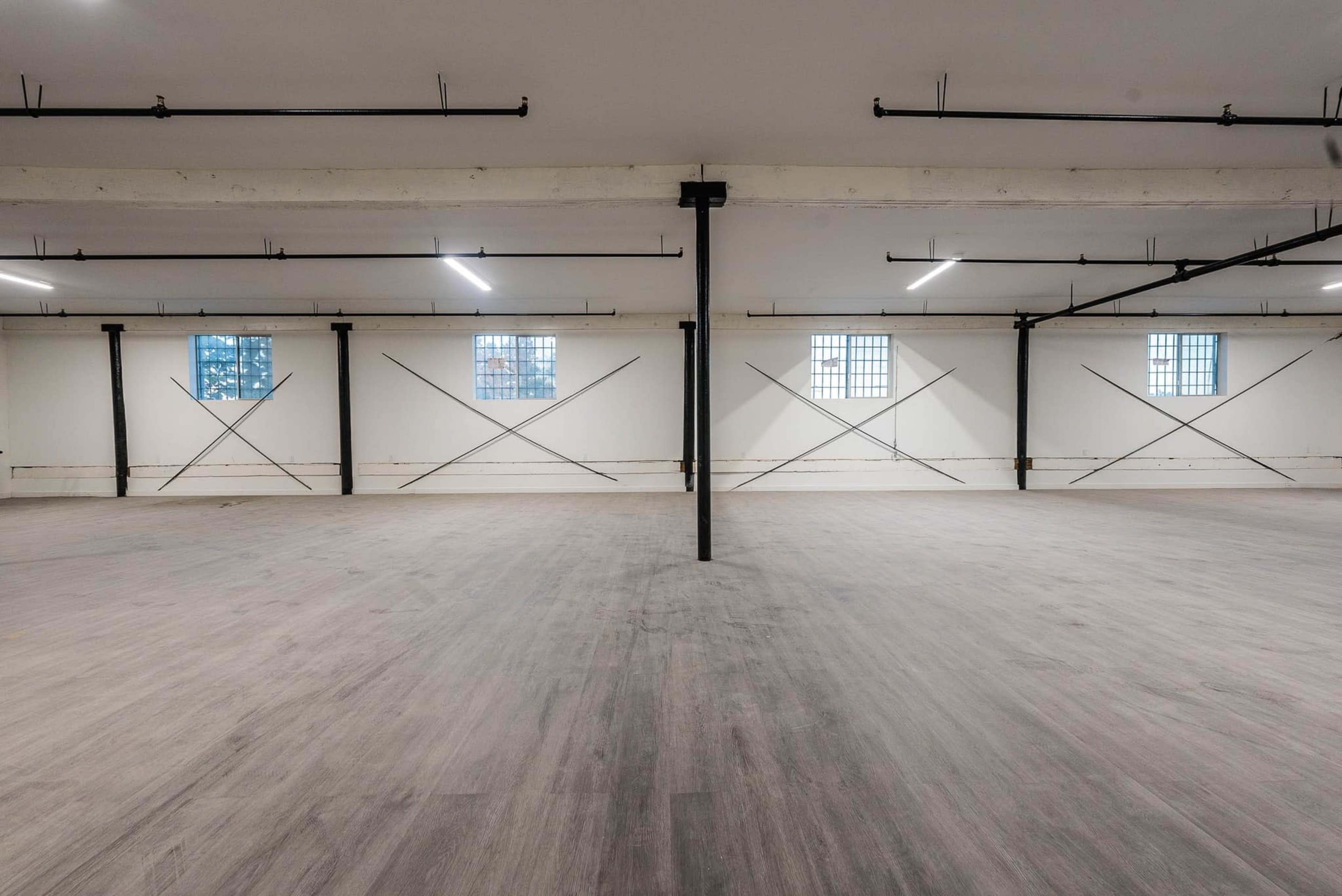 The image shows a spacious, empty industrial-style room with large windows and exposed structural elements.