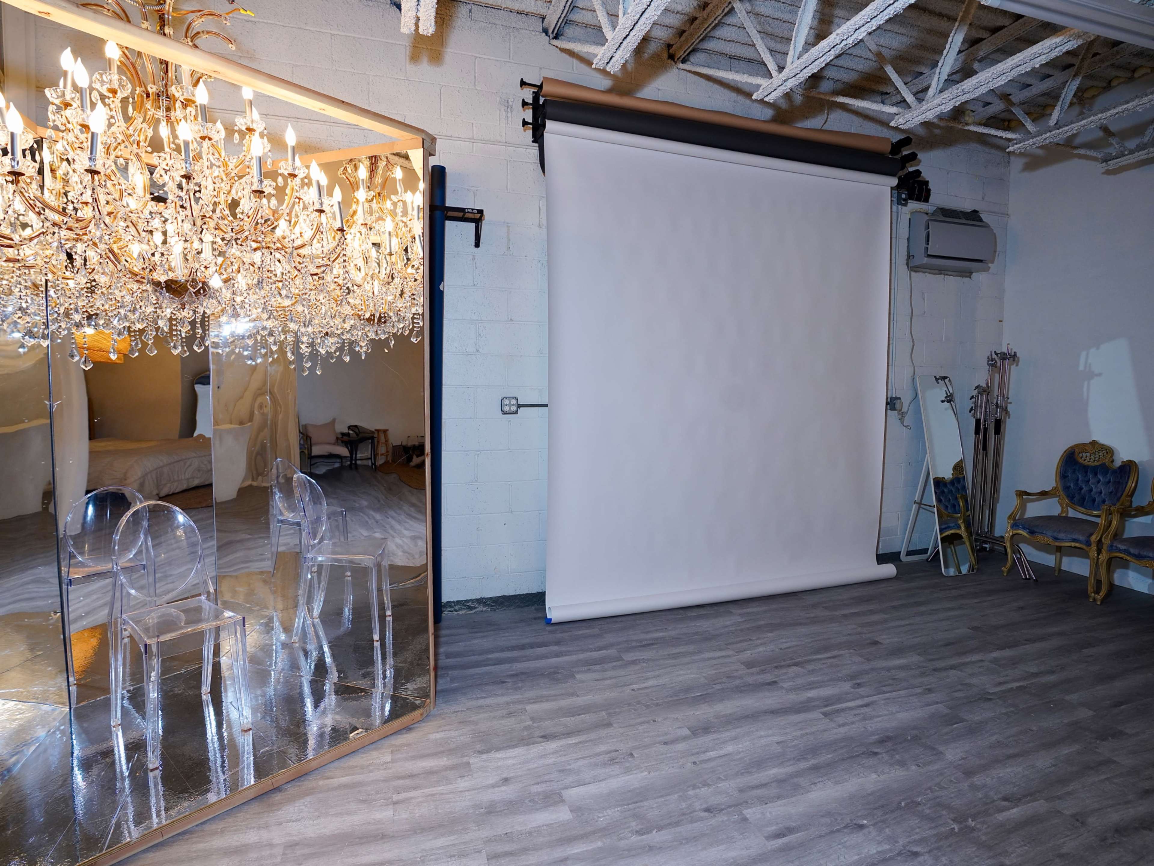 The image shows a photography studio featuring a large mirror with a chandelier, a blank white backdrop, and a decorative chair in the corner.