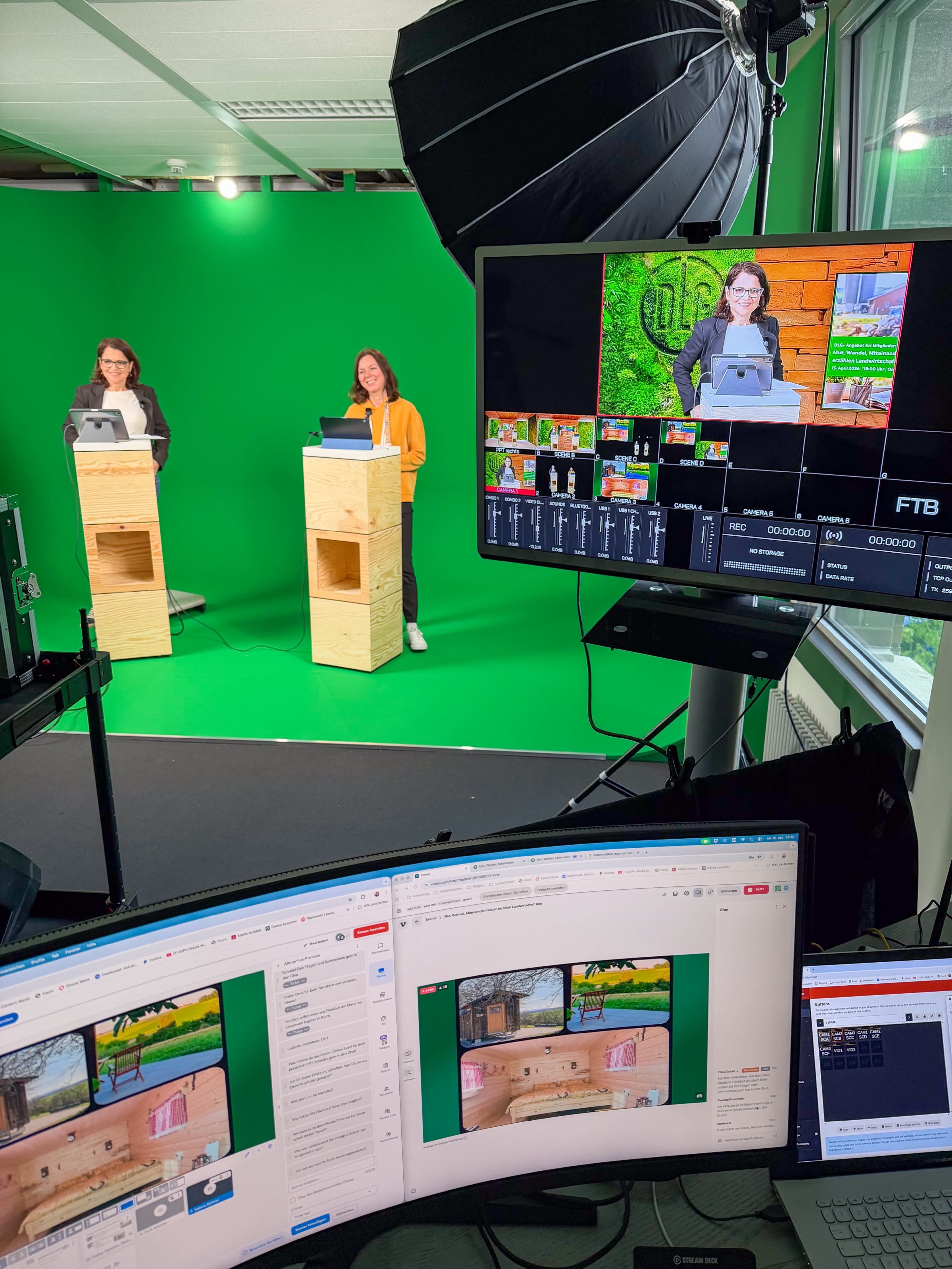 Two women are standing behind wooden podiums in front of a green screen, while a computer monitor displays a broadcast setup with multiple camera feeds.