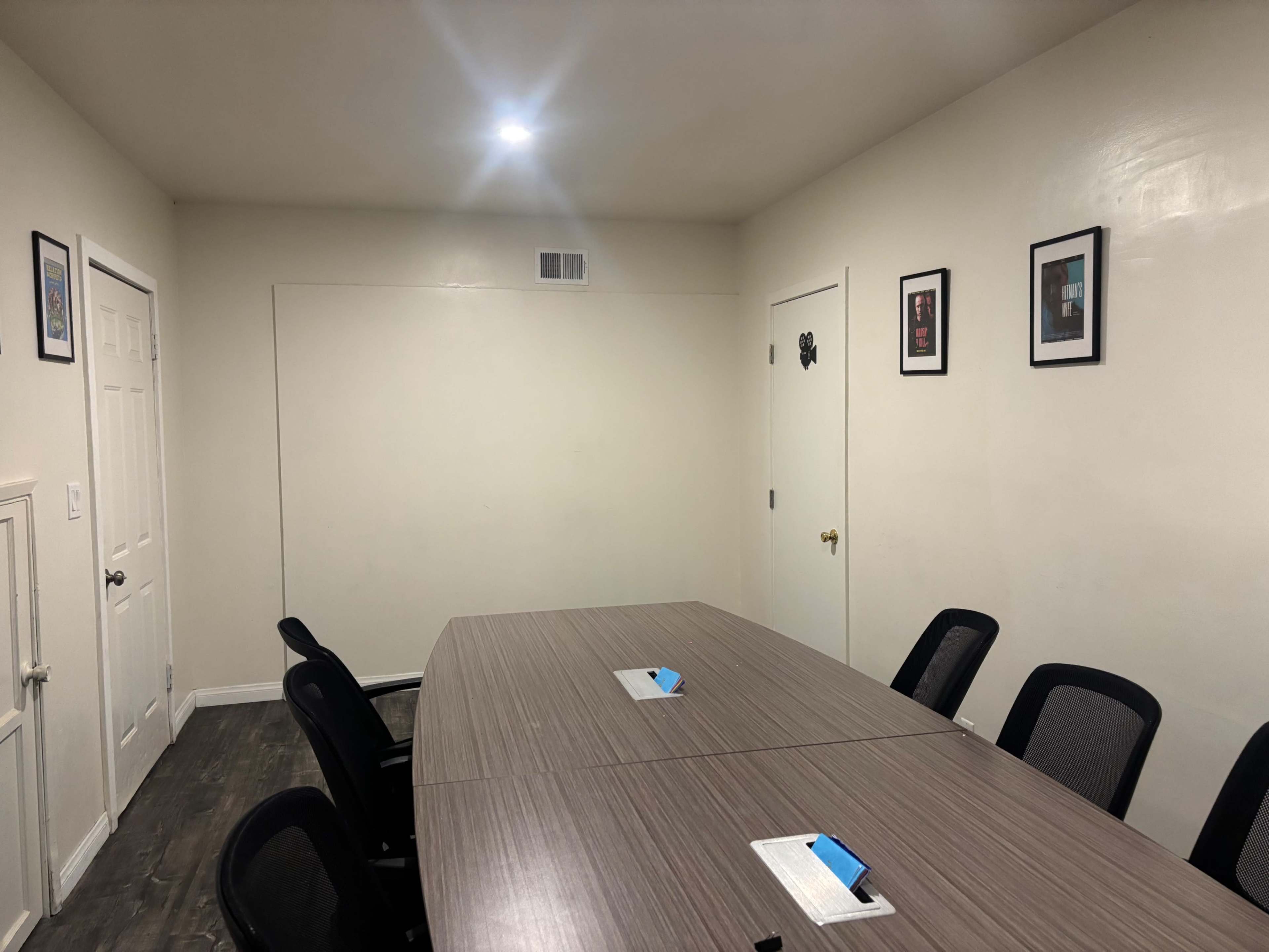 A conference room features a large oval table surrounded by black chairs, with framed artwork on the walls and a door leading to another area.