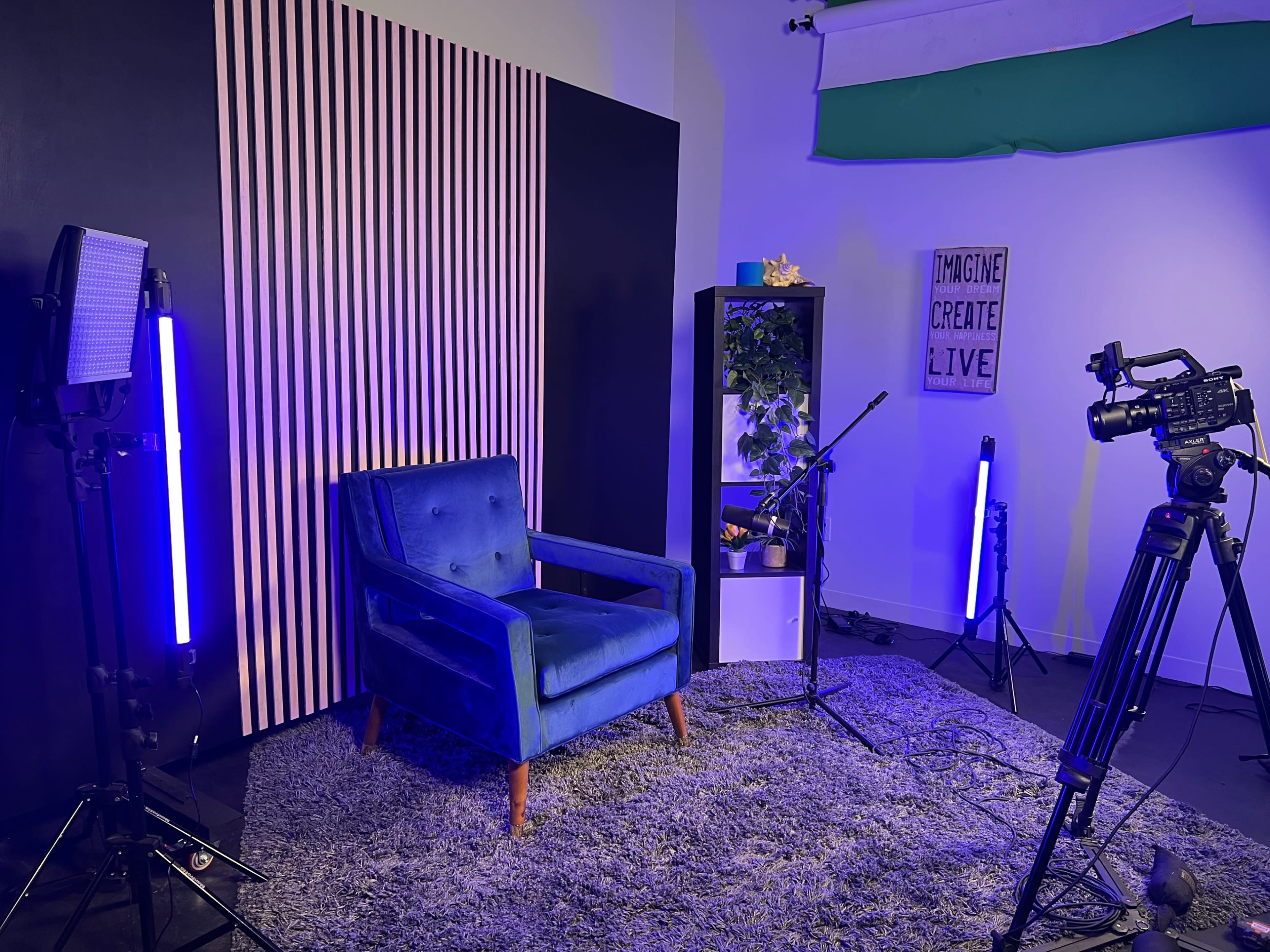 A studio setup with a blue armchair, a backdrop of striped panels, a plant, and two lights beside a camera on a tripod.