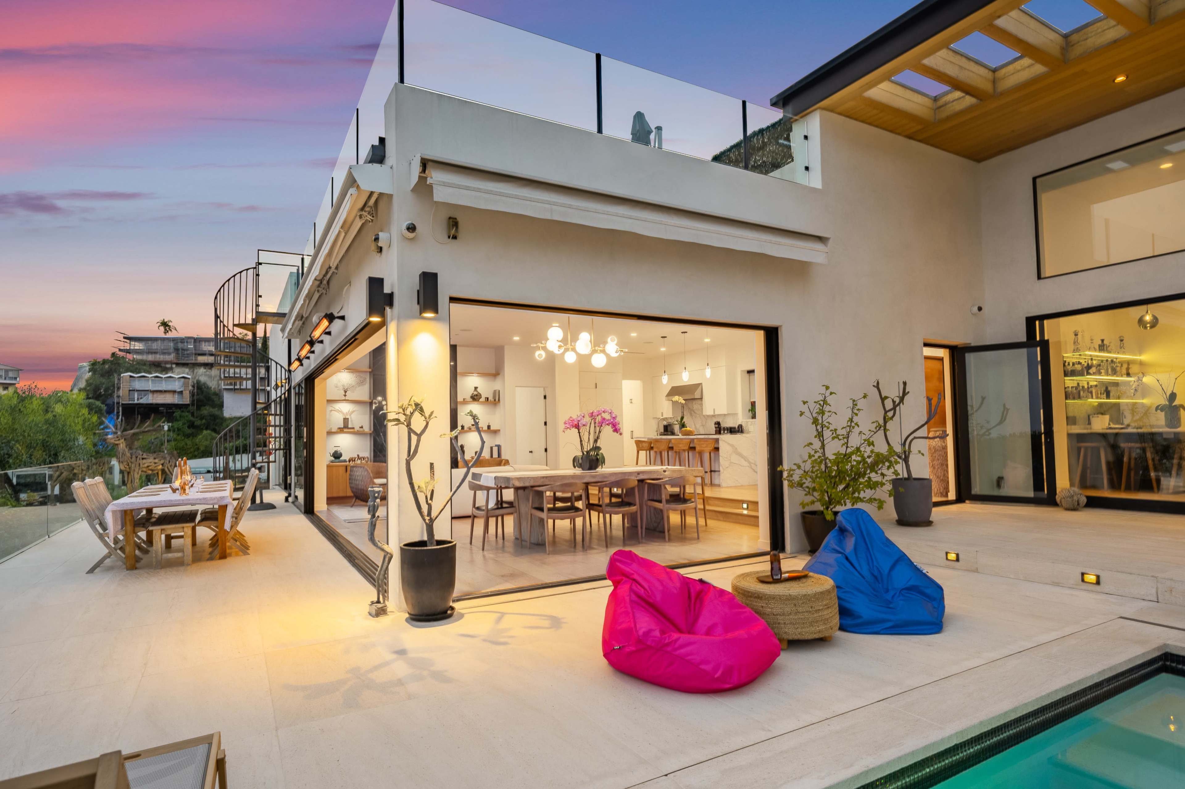 The image shows a modern house with large glass doors opening to a patio featuring bean bag chairs and a pool.