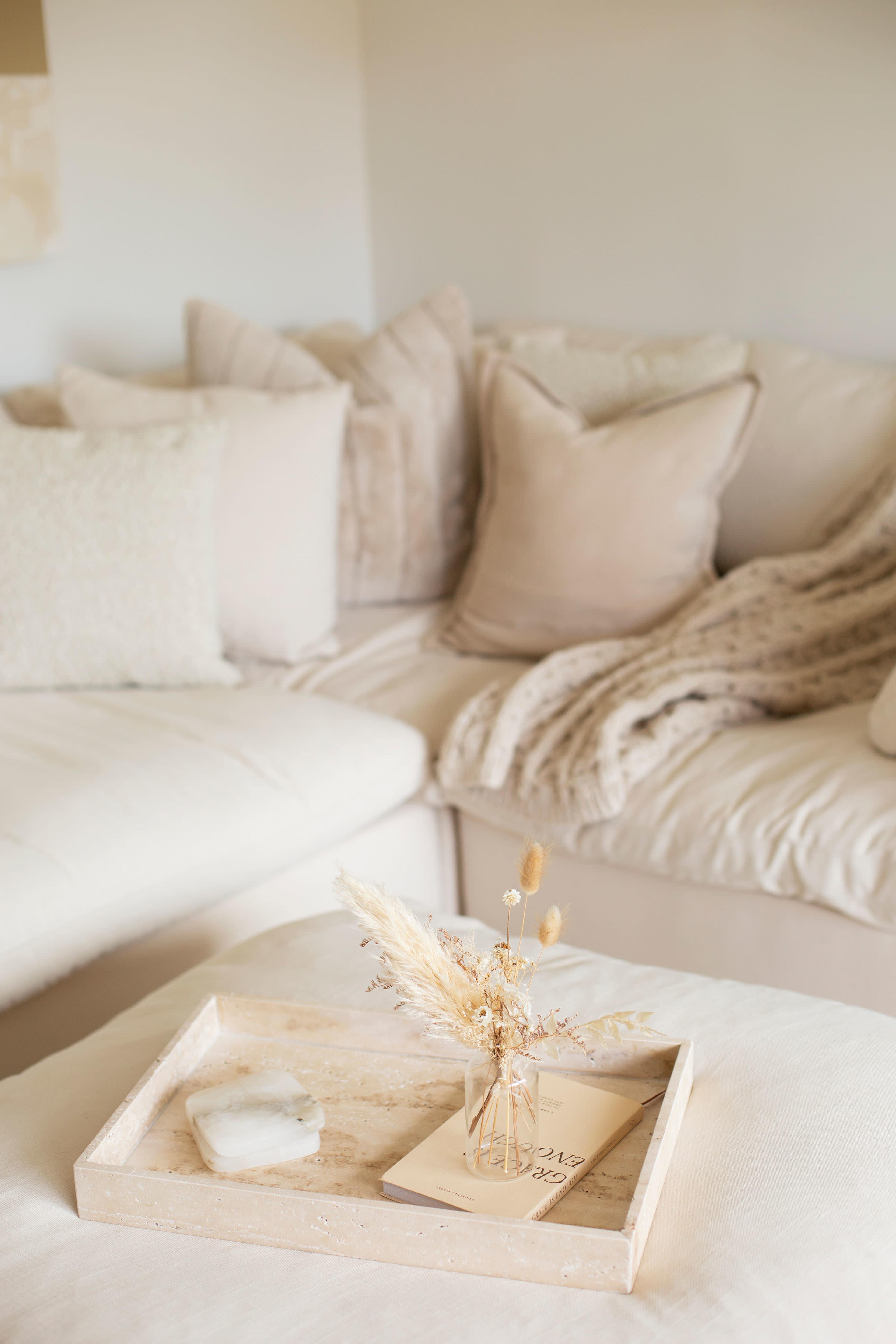 A cozy living room features a light-colored sectional sofa adorned with numerous cushions, alongside a wooden tray holding a small vase and a book.