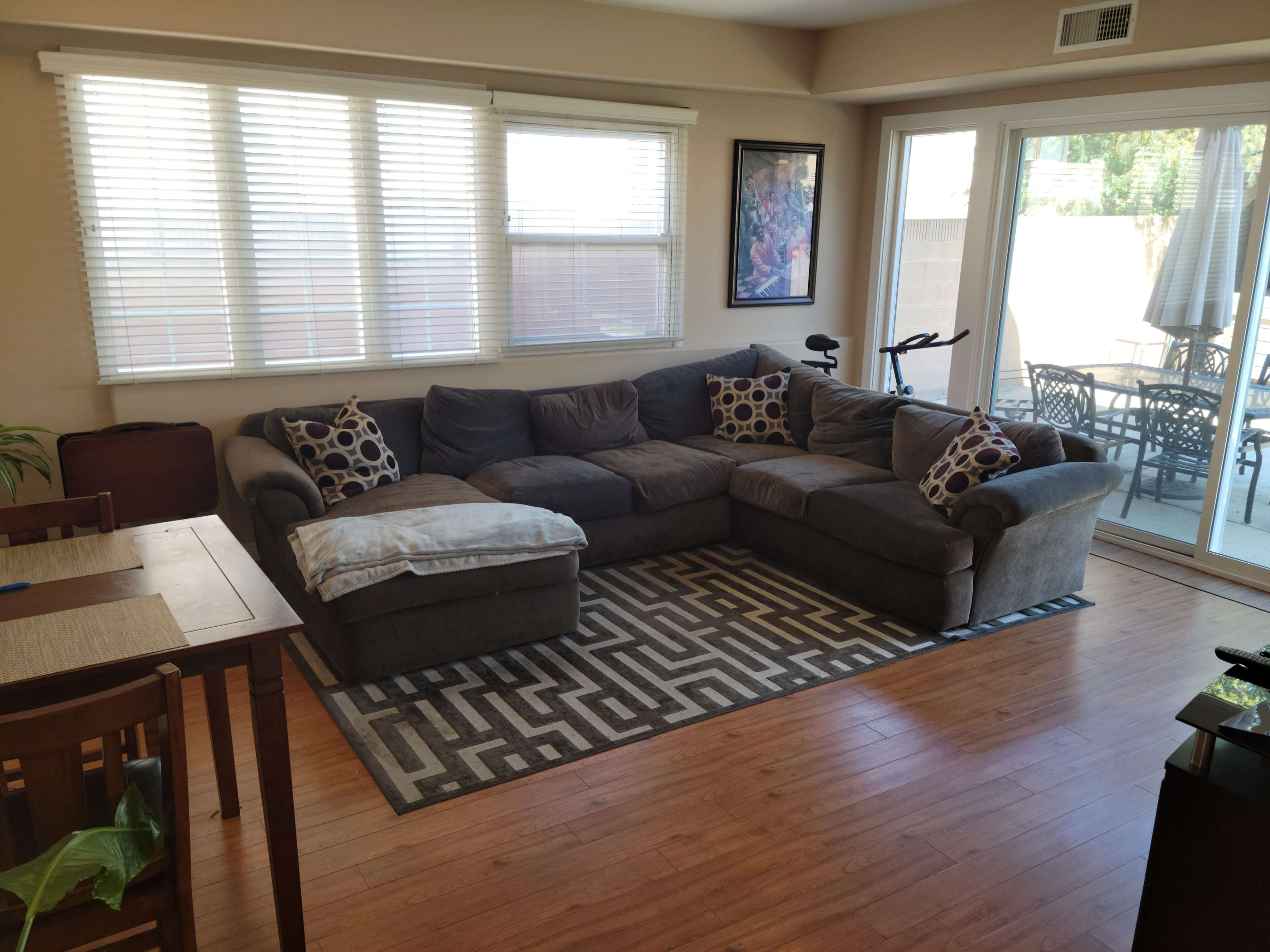 The living room features a spacious sectional sofa with patterned cushions, a wooden dining table, and large windows that provide natural light.
