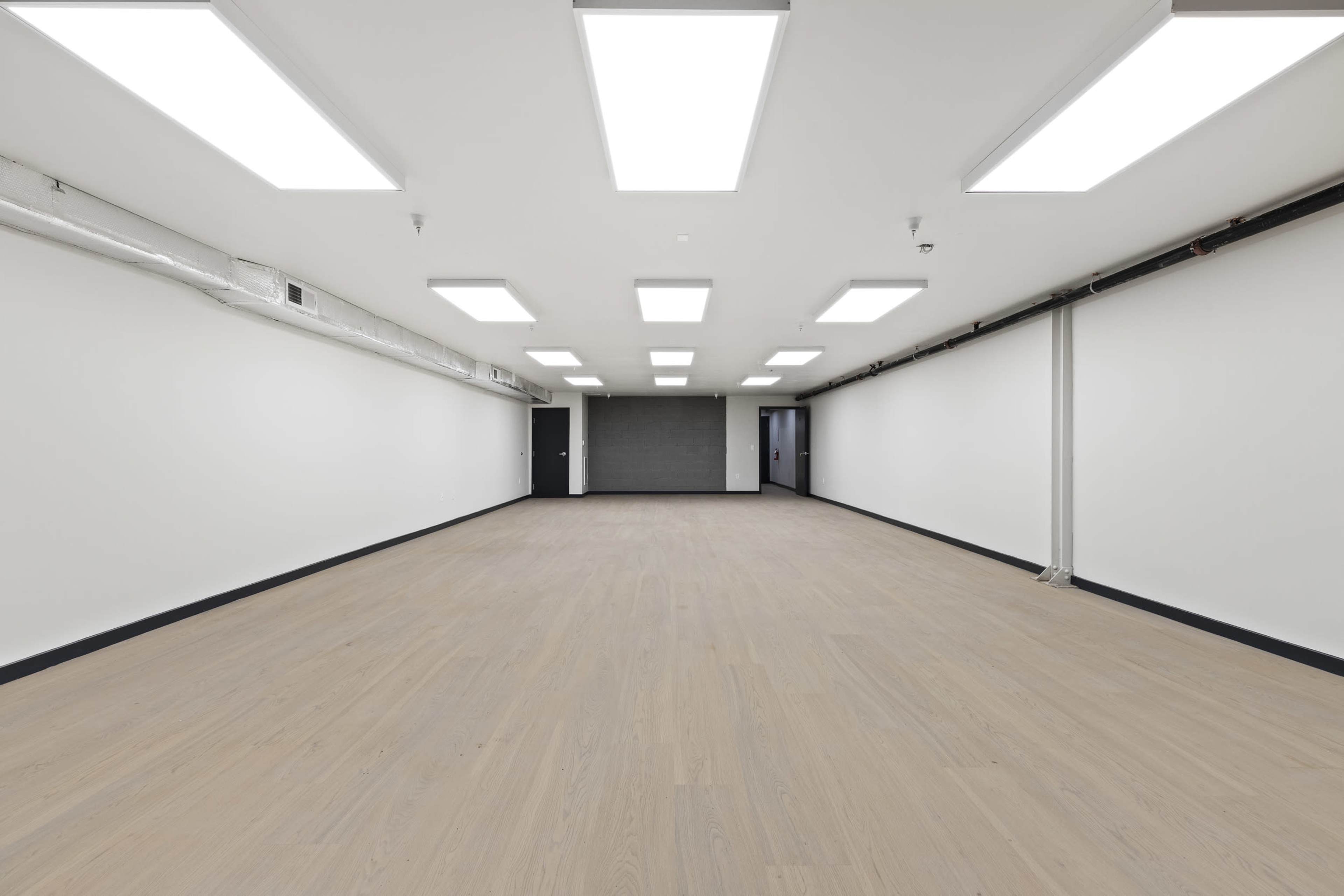 The image shows a large, empty room with light wooden flooring, white walls, and bright, overhead lighting.