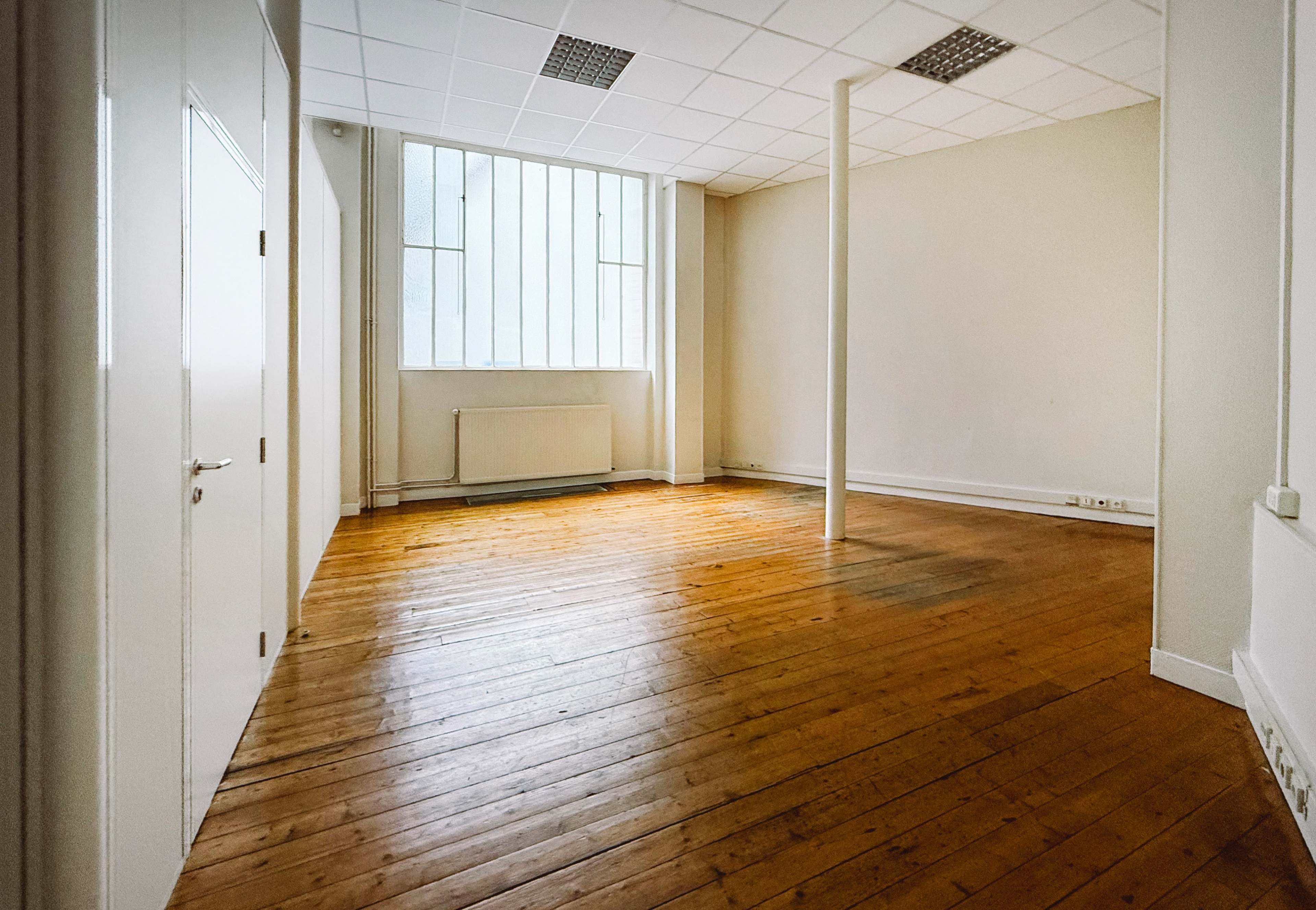 The image shows a vacant room with wooden floors, white walls, and large windows allowing natural light to enter.