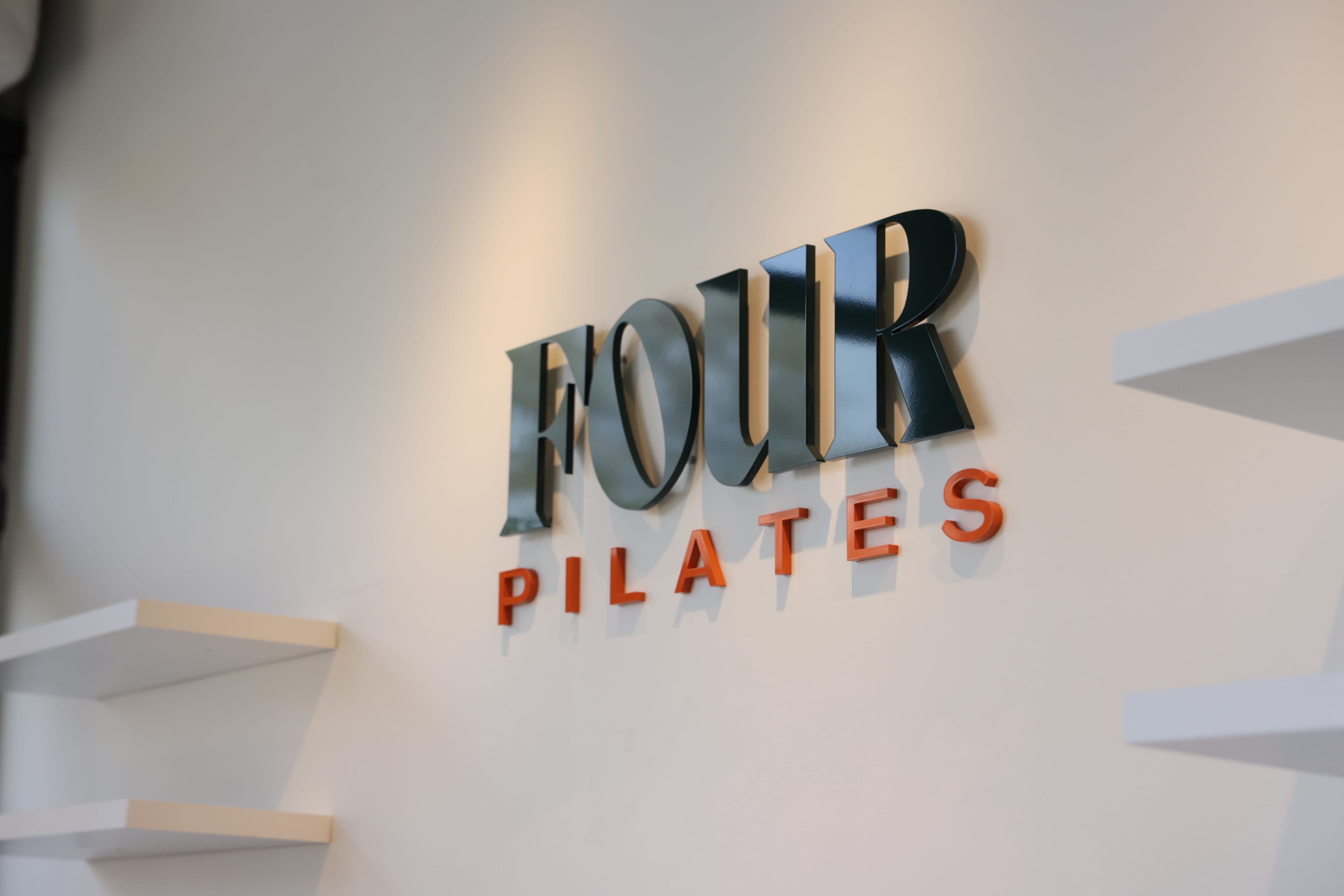 The image shows a wall featuring the sign "FOUR PILATES" in a three-dimensional design, with the word "FOUR" in dark colors and "PILATES" in orange.