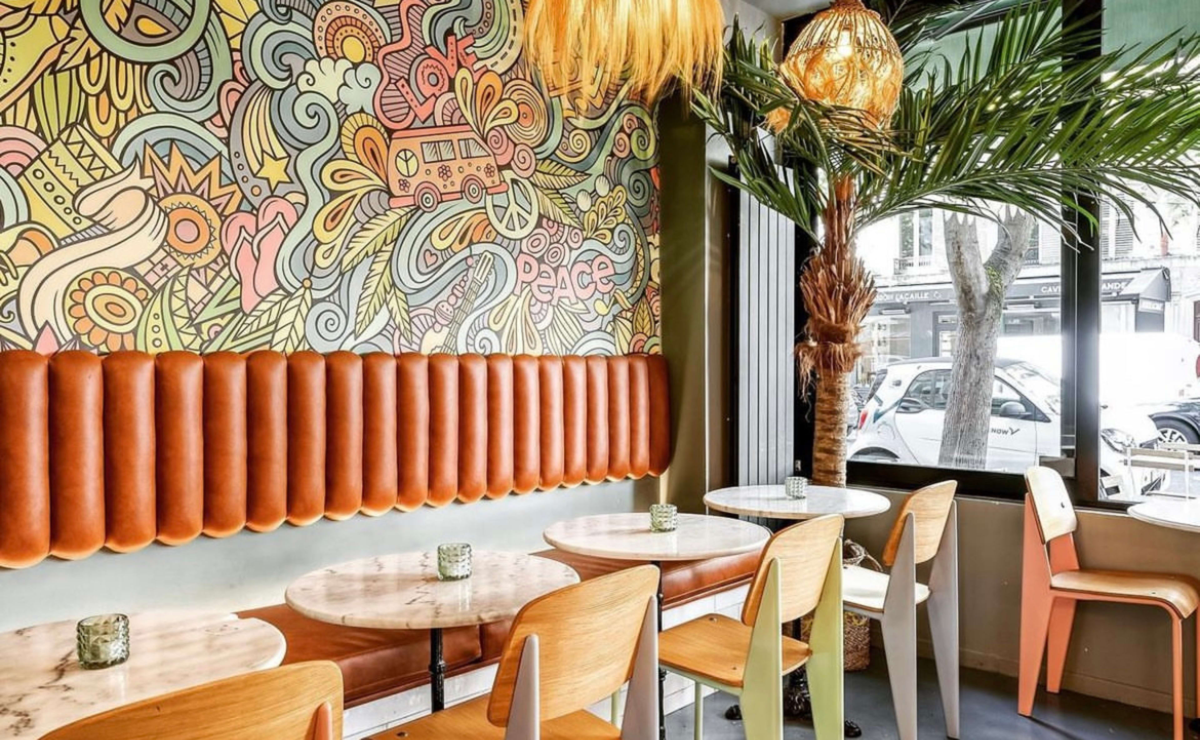 A brightly decorated café interior featuring a colorful mural, palm fronds, and round tables with wooden chairs.