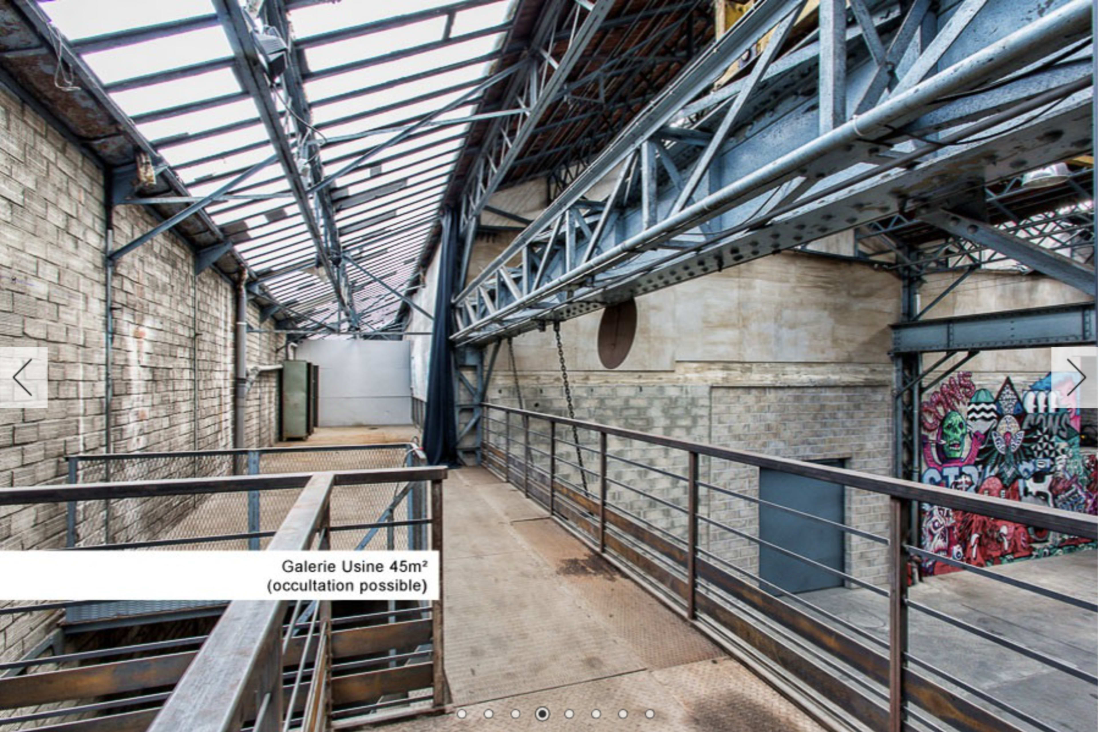 An industrial-style gallery with metal beams and a graffiti-covered wall, featuring a walkway overlooking a spacious area below.