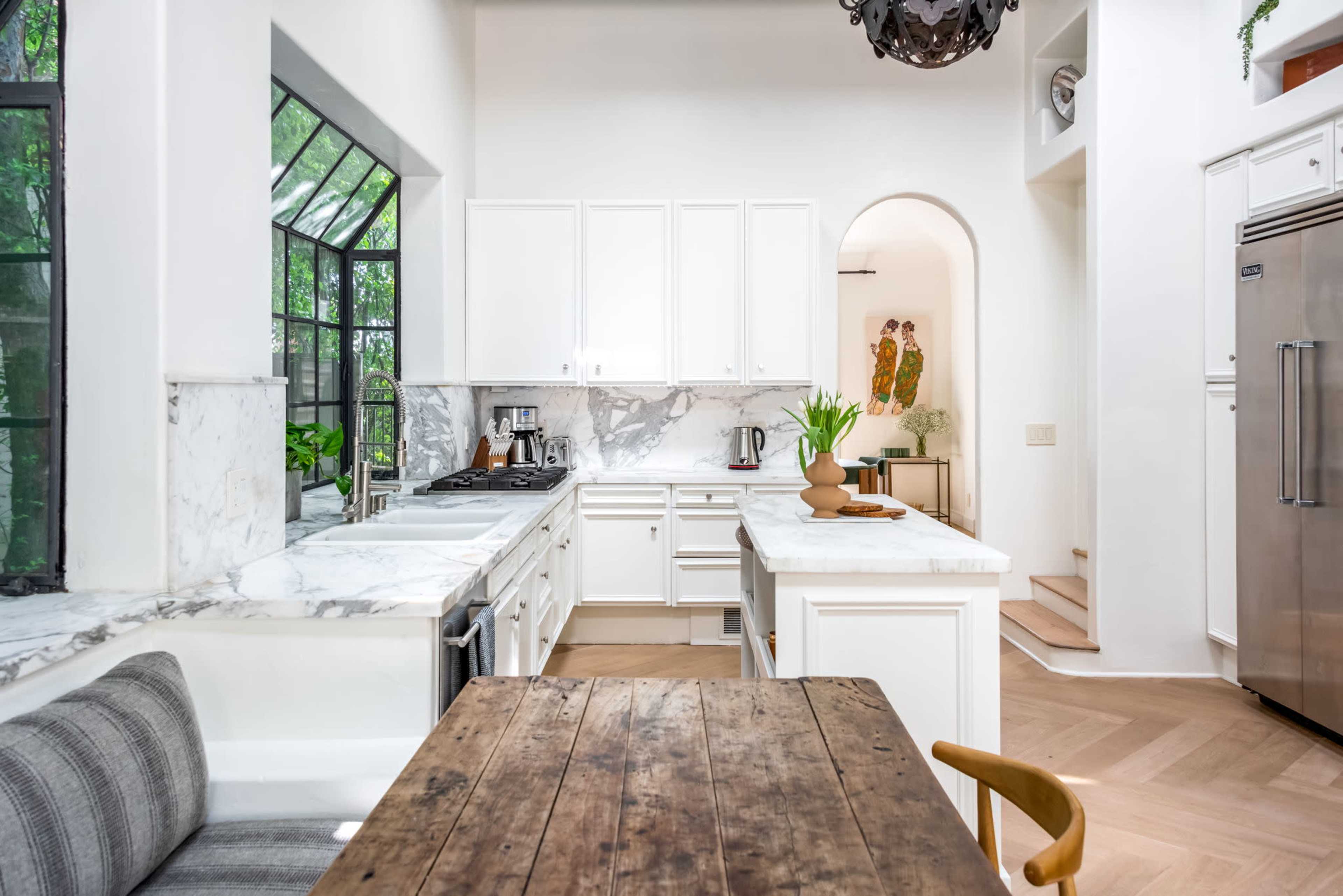 The image shows a bright kitchen with marble countertops, white cabinetry, and a dining area featuring a wooden table and a bench.