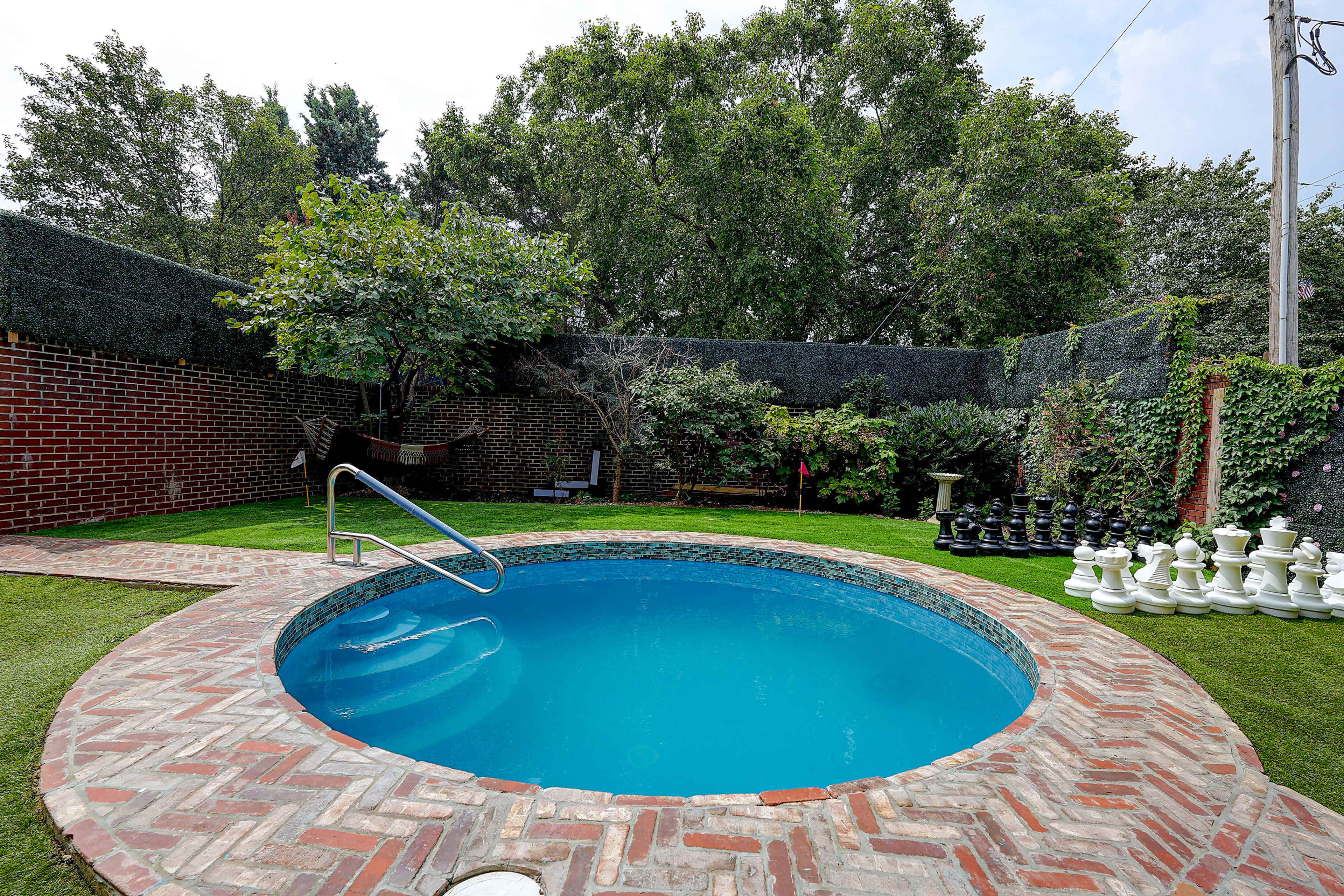 A circular pool is set in a brick patio surrounded by lush greenery and a large chess set on a lawn.