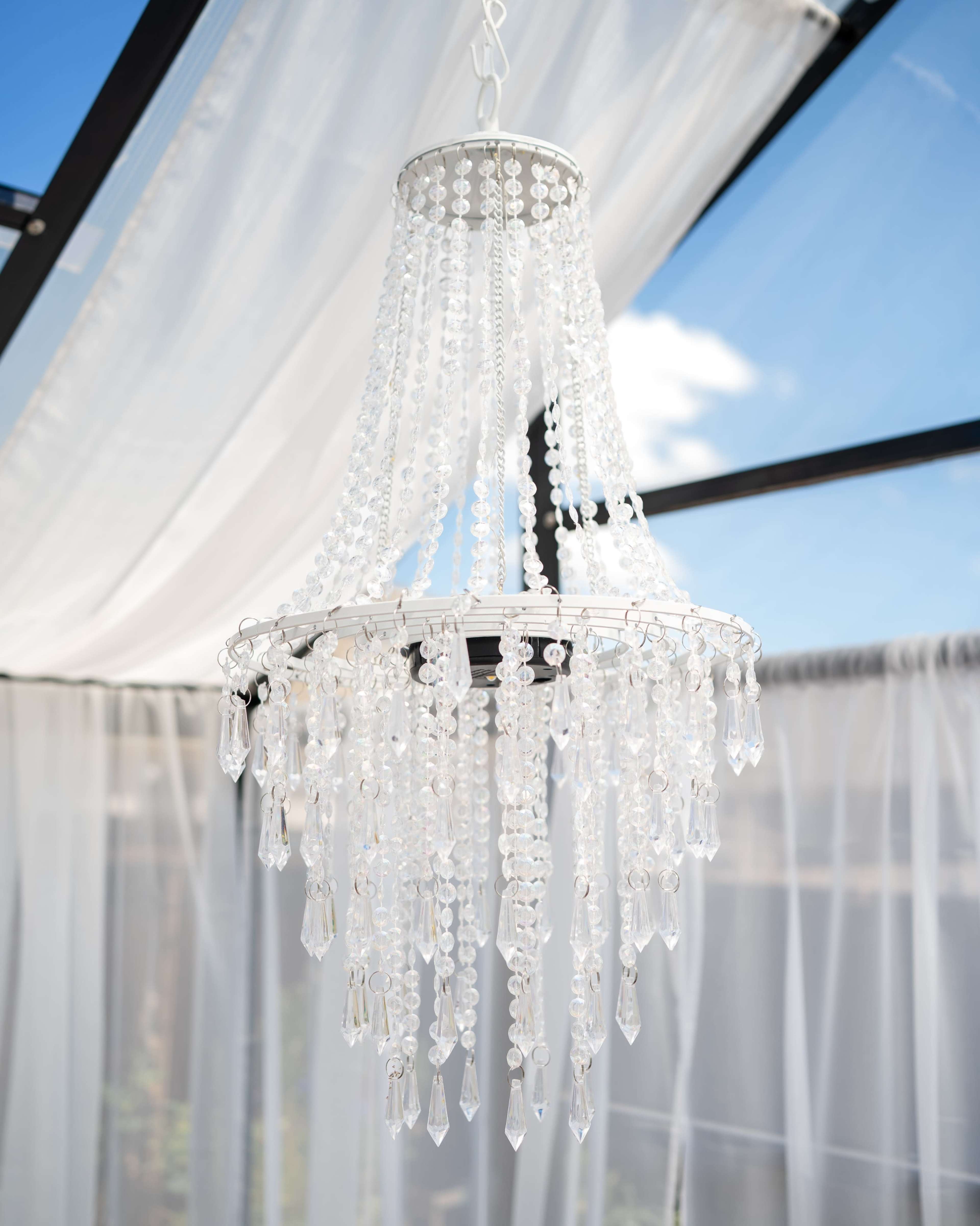 A white chandelier adorned with transparent beads and crystals hangs from a ceiling beneath a blue sky visible through a glass roof.