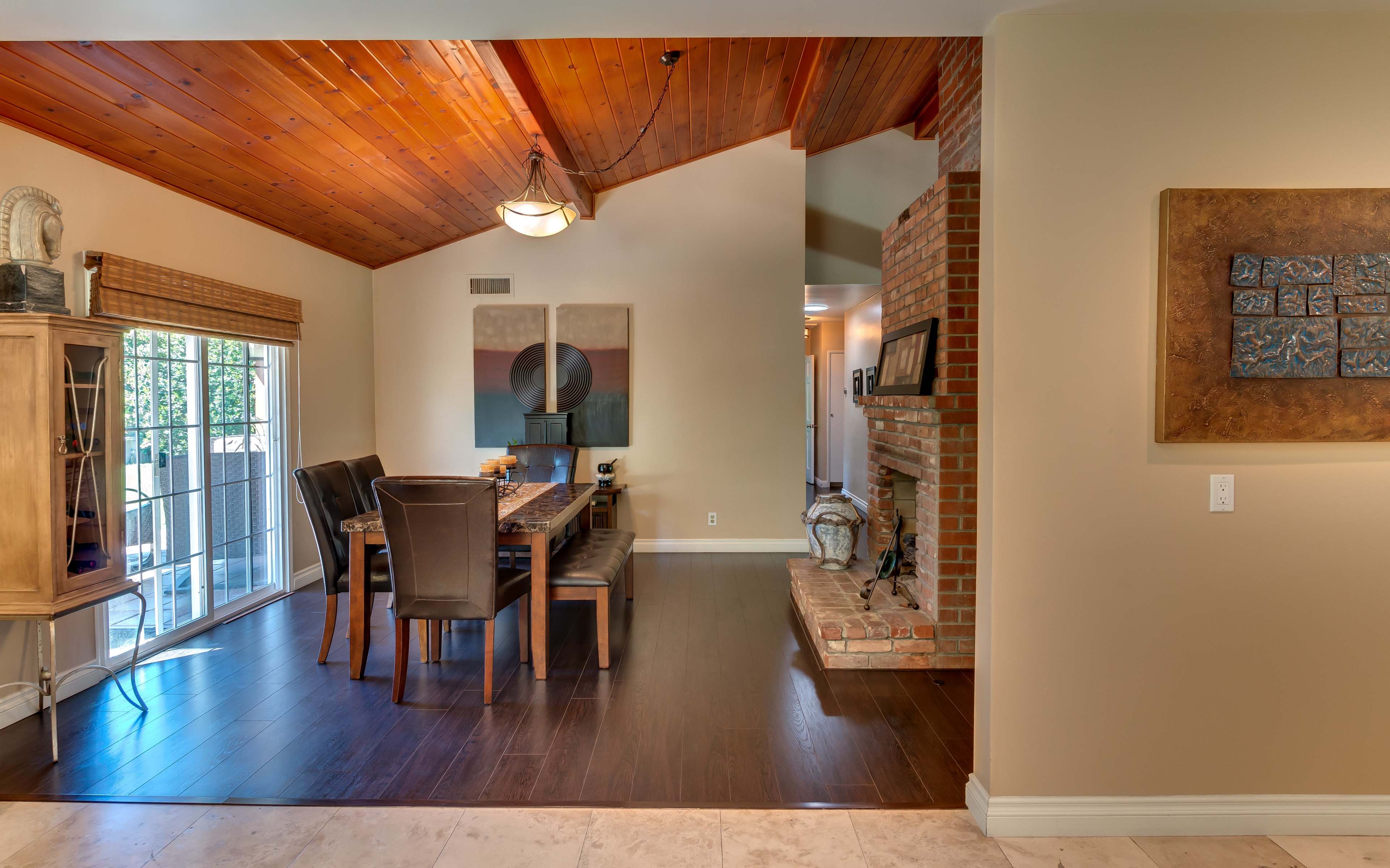 A dining area with a wooden table and chairs, a brick fireplace, and large windows allowing natural light to enter.