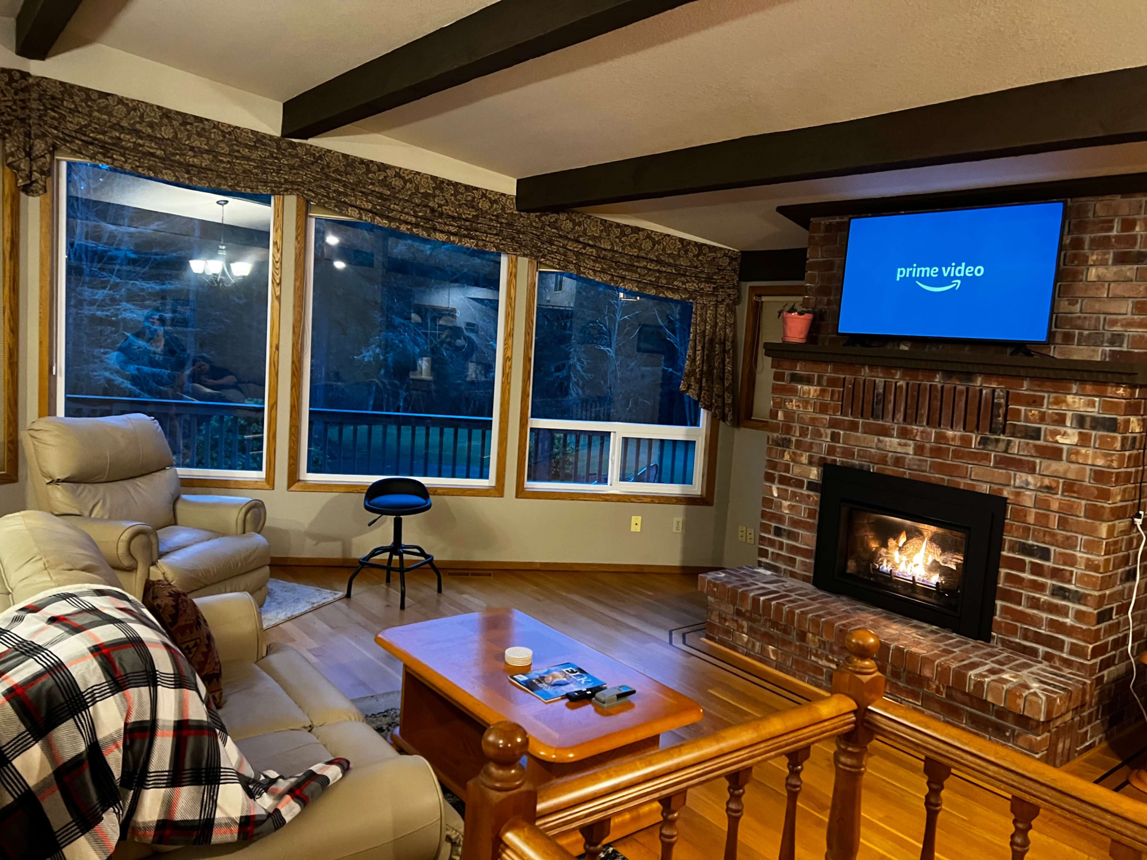 A cozy living room features a fireplace, a large window with a view, and a television displaying the Prime Video logo.
