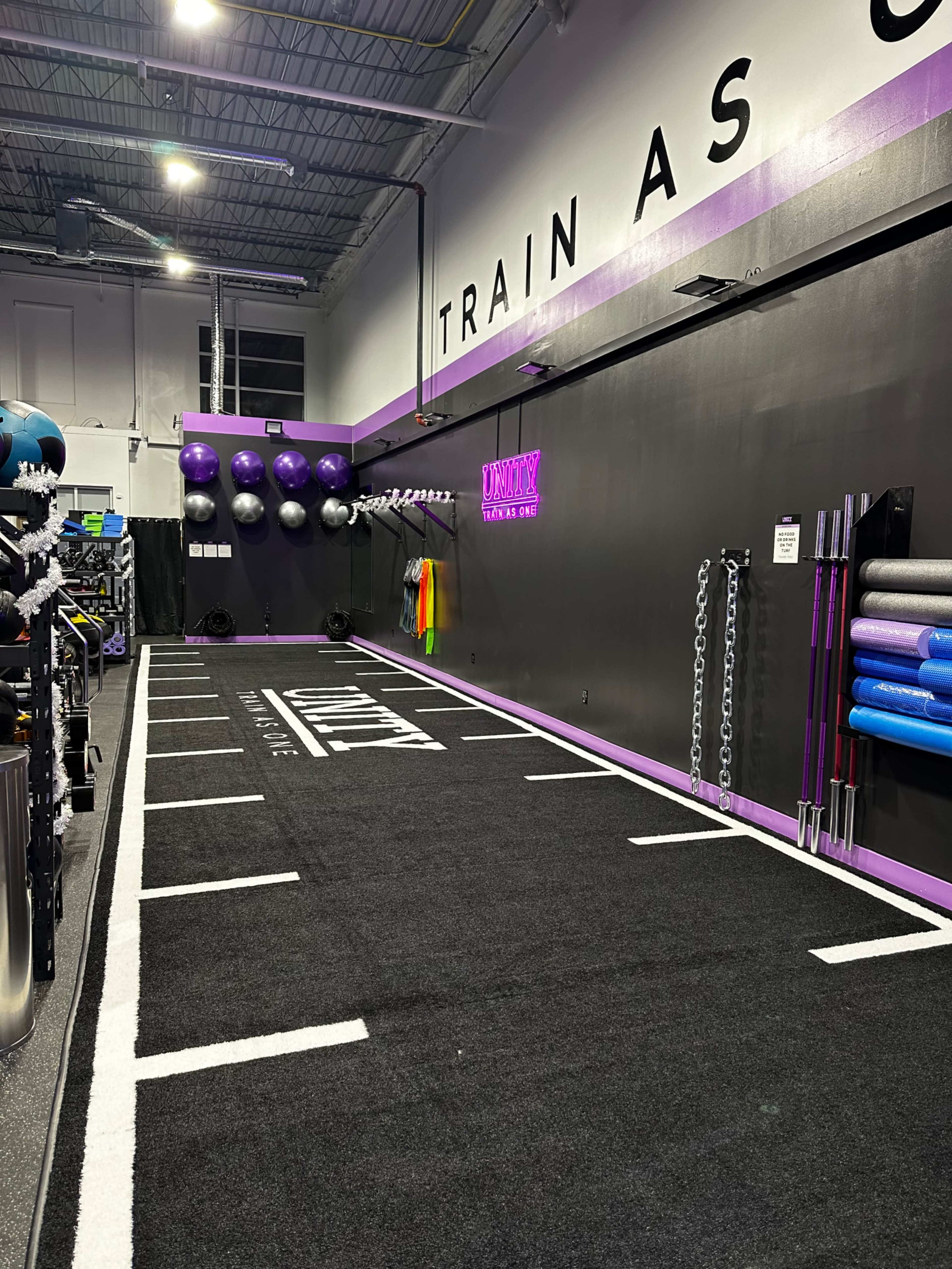 A fitness training area with a black turf floor marked by white lines, featuring exercise equipment and a wall adorned with fitness balls and foam rollers.