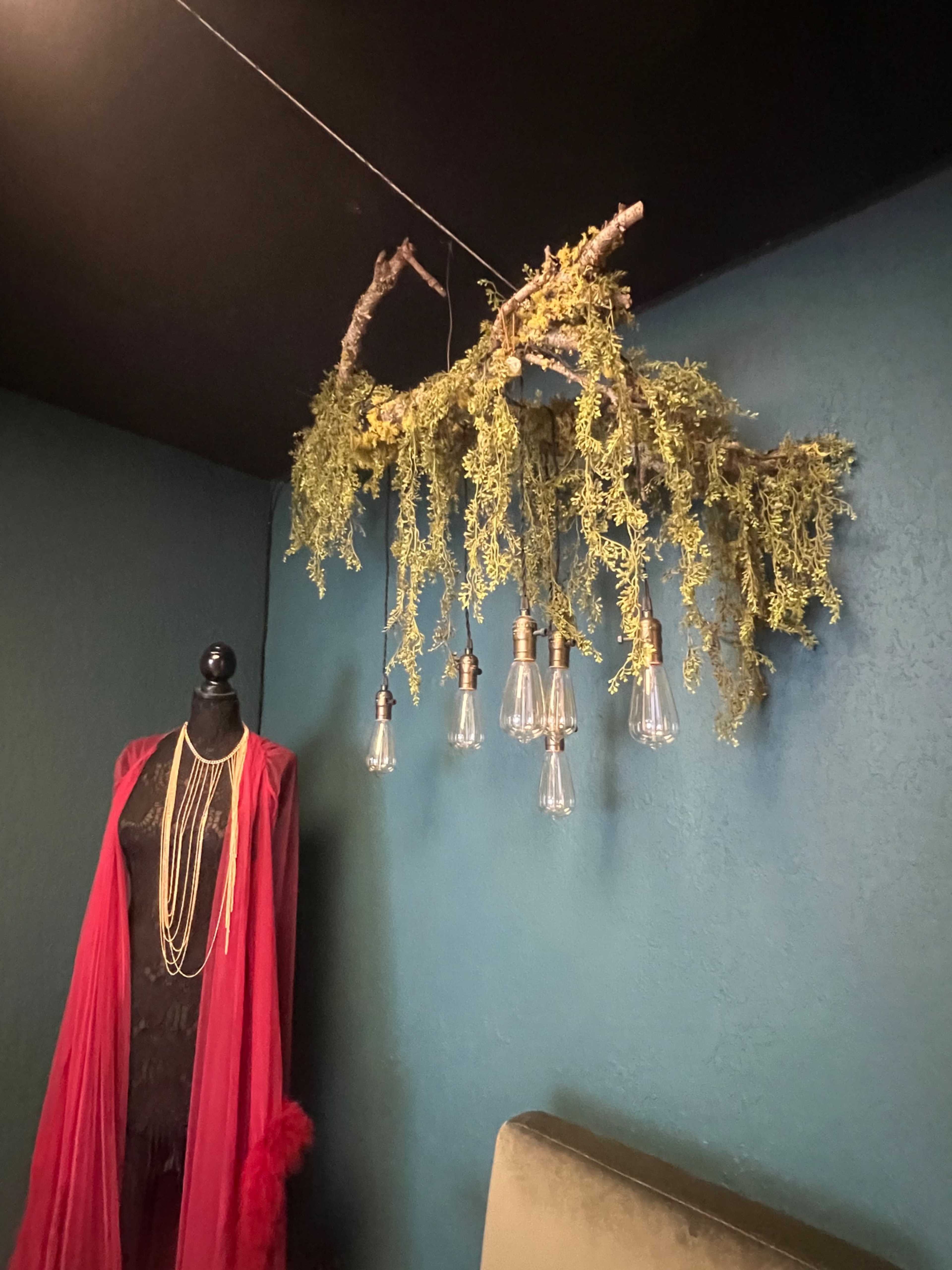 A branch adorned with greenery and pendant light bulbs hangs from the ceiling above a mannequin dressed in a black outfit and draped with a red shawl.