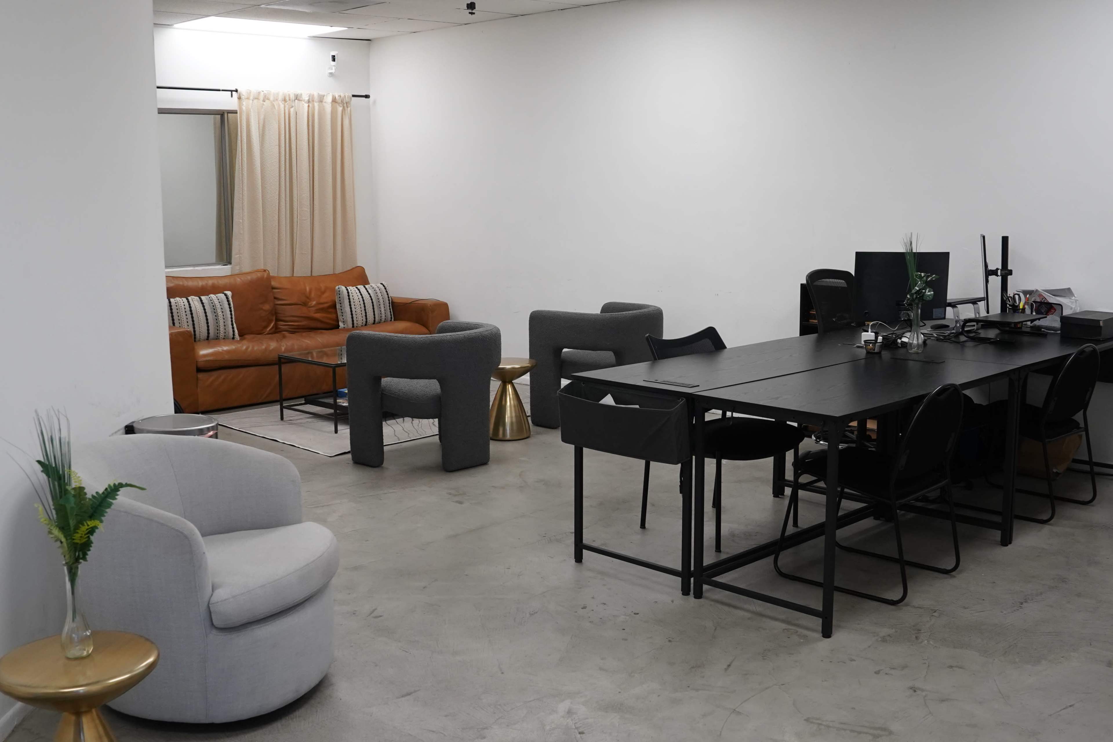 A modern office space with a seating area featuring a brown leather couch and gray armchairs, alongside a black rectangular table surrounded by several chairs and a desk with office equipment.