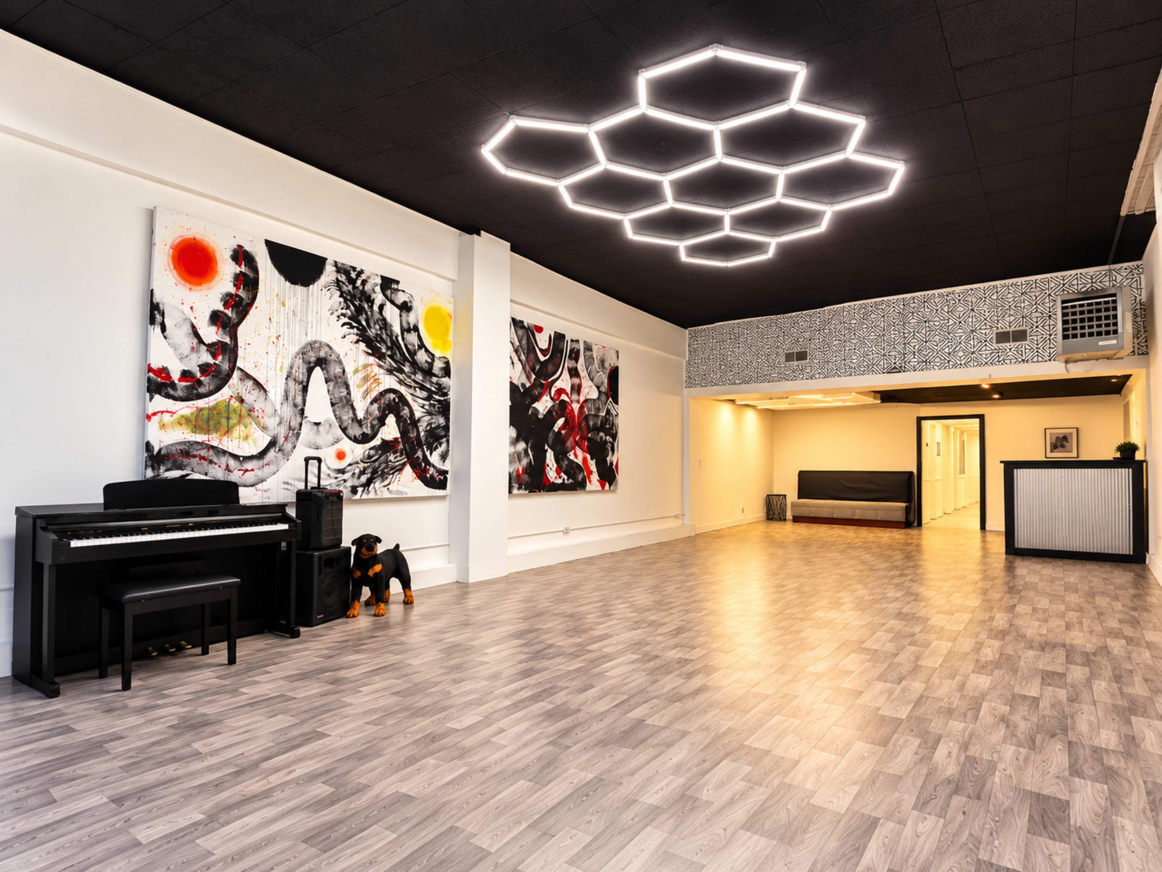 The image shows a spacious dance studio with light-colored wooden flooring, black ceiling tiles, modern hexagonal lighting, abstract artwork on the walls, and a piano positioned in one corner.
