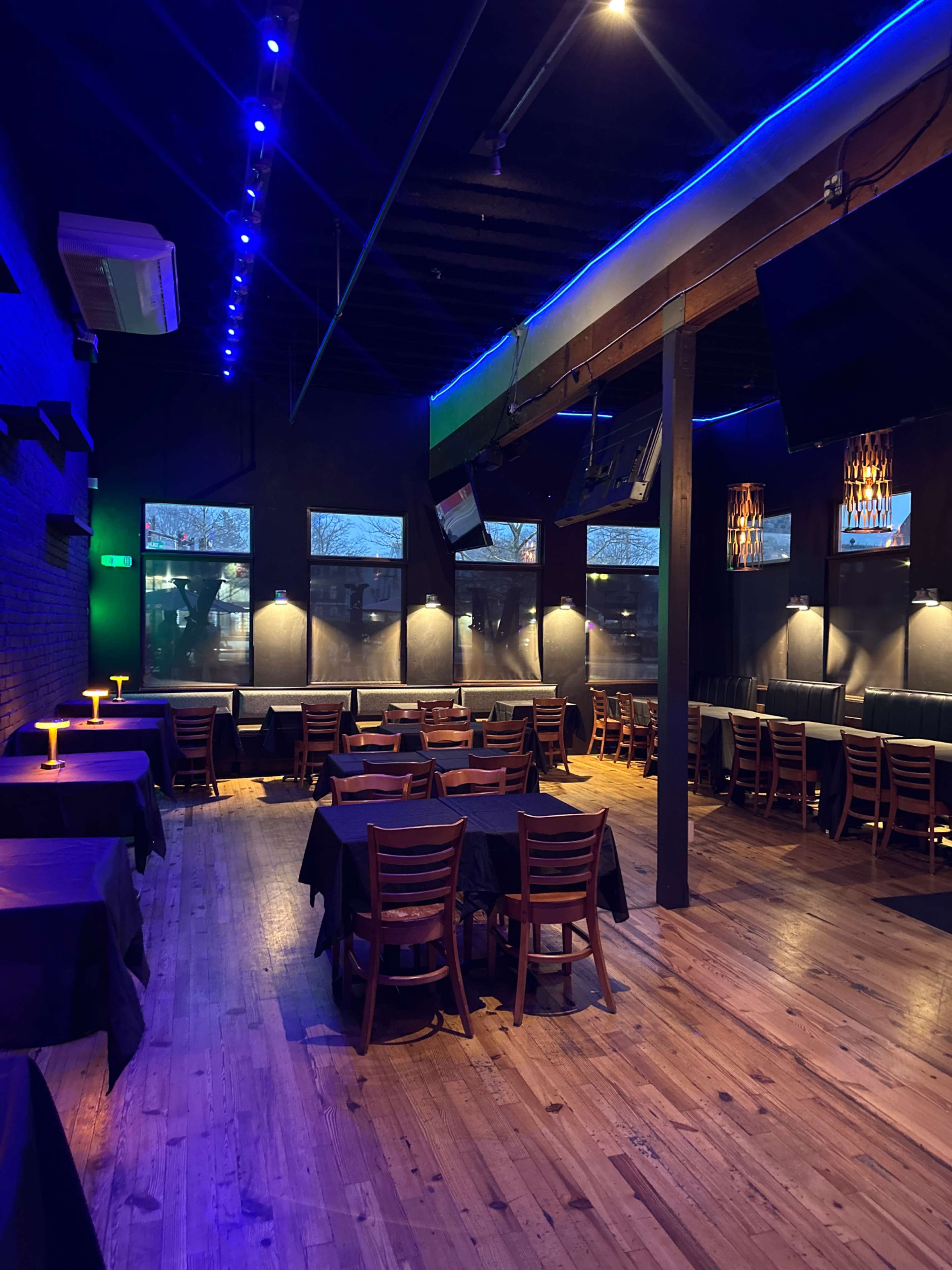 A dimly lit restaurant interior with wooden floors, black tablecloths on tables, and blue accent lighting.