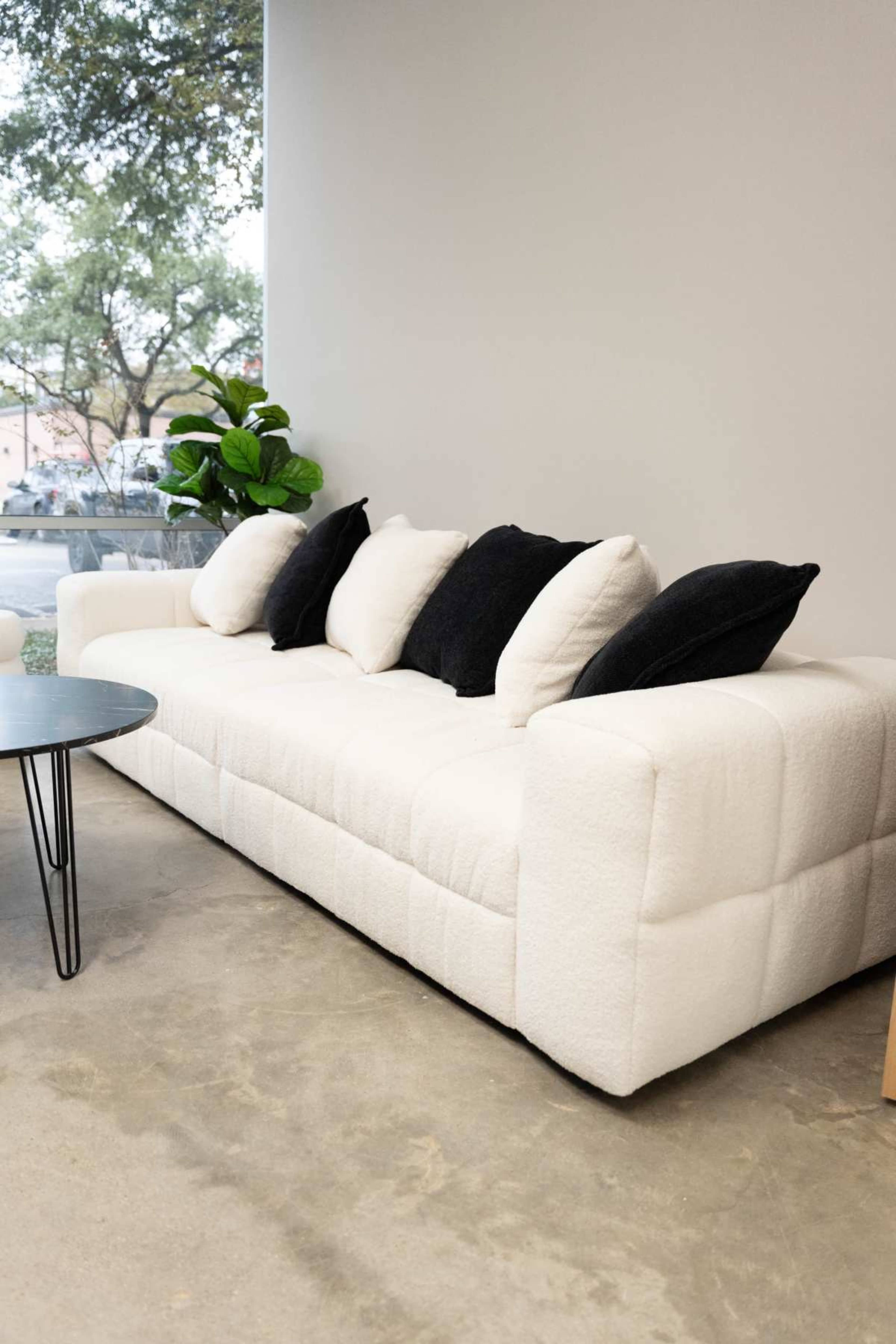 A modern white sofa with four cushions sits against a wall next to a small black table and a large window.