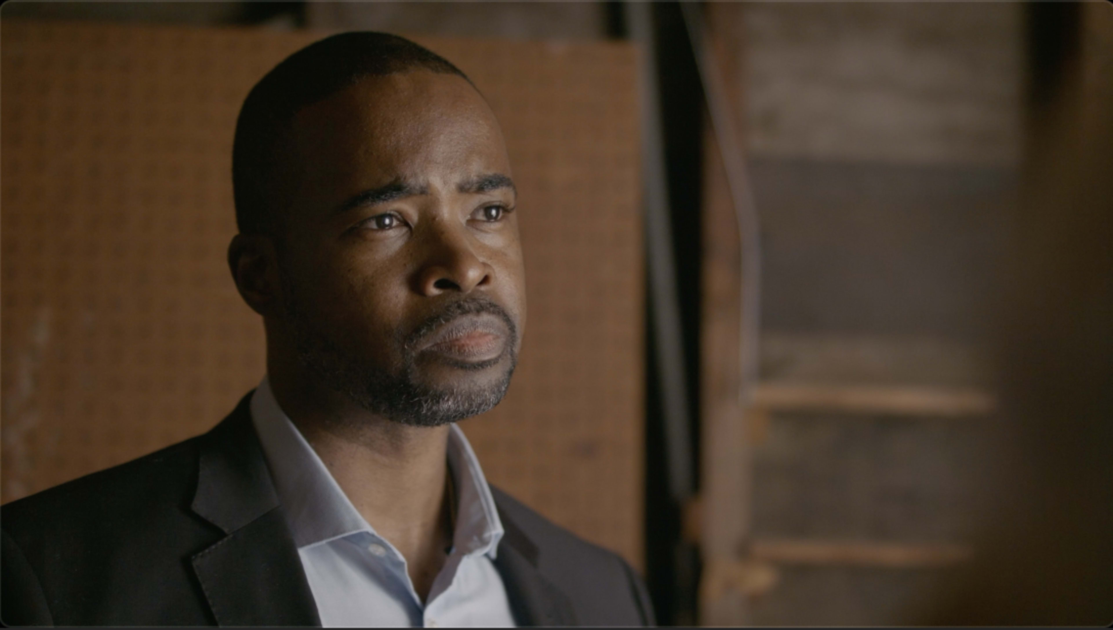 A man in a suit stands in a dimly lit environment, looking intently off to the side.