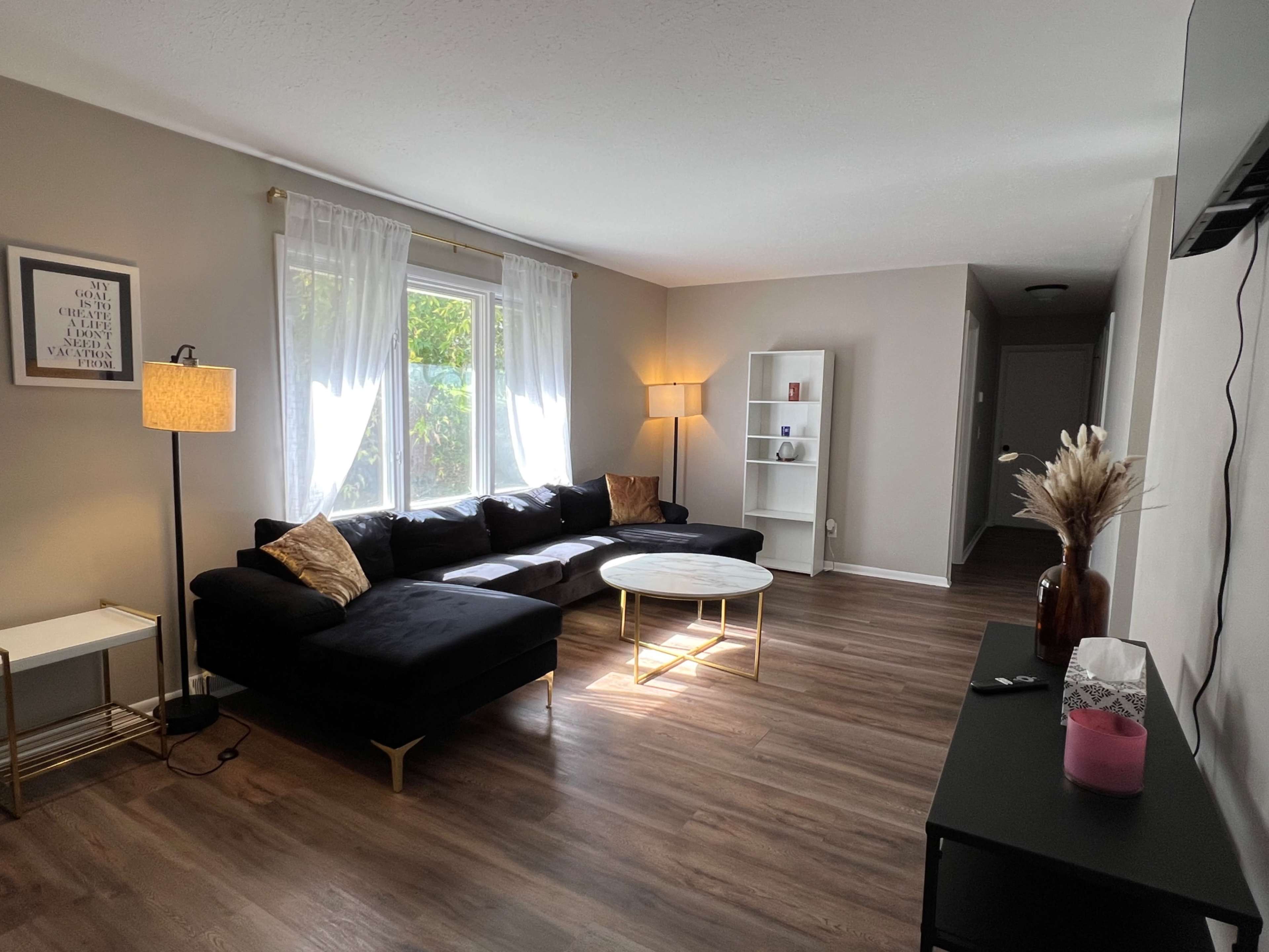 The living room features a dark sectional sofa, a circular coffee table, large windows with sheer curtains, and a floor lamp, all set on hardwood flooring.