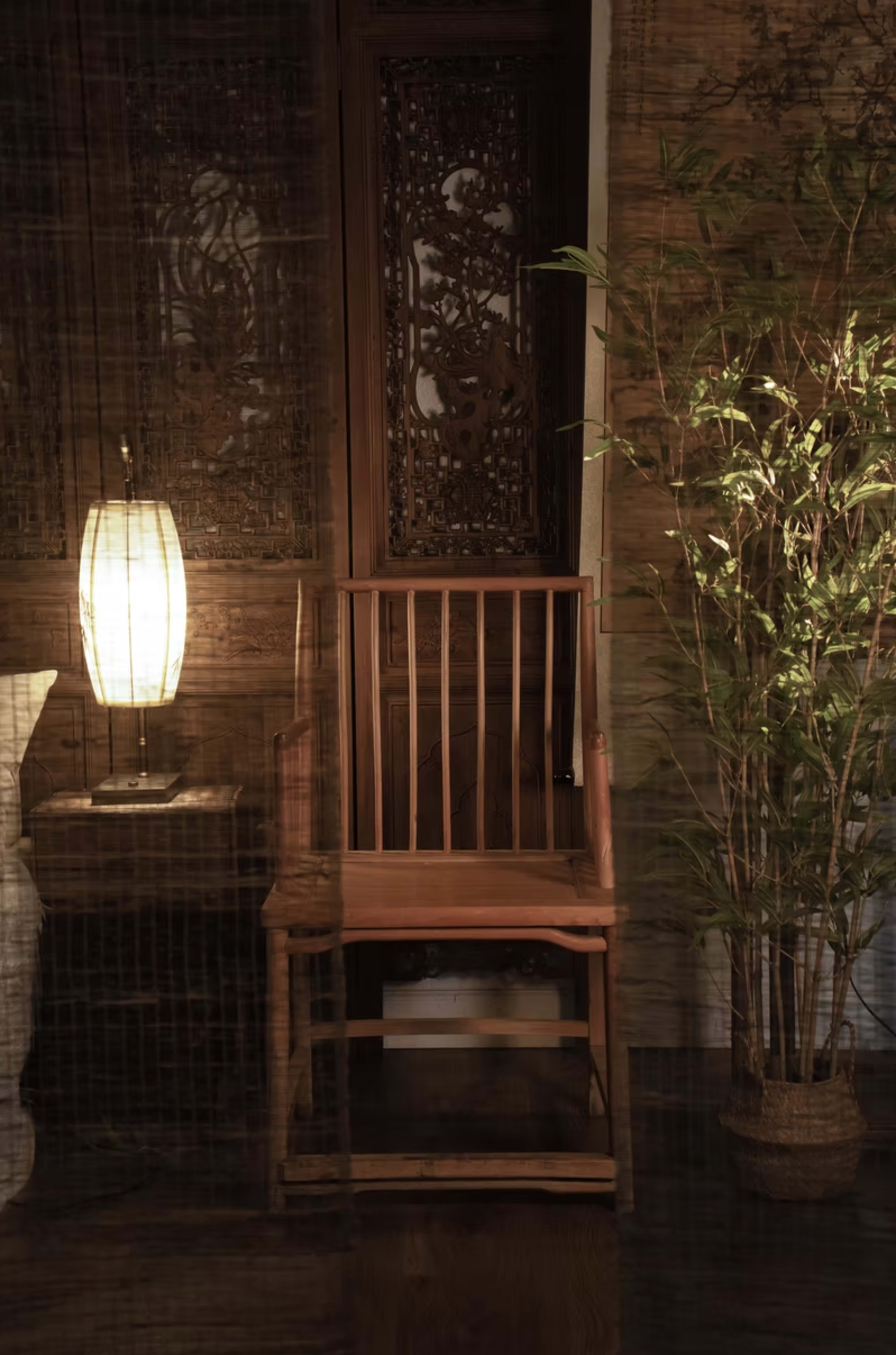 The image shows a wooden chair next to a lamp and a potted plant in a softly lit interior space.