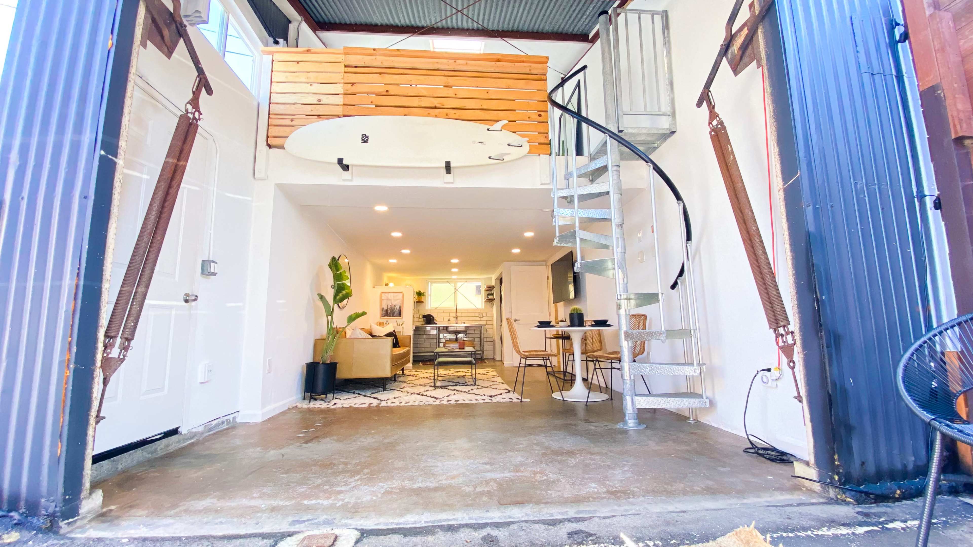 The interior entrance of a modern space featuring a spiral staircase leading to a loft area, accompanied by a surfboard mounted on the wall and a lounge area with a couch and dining table.