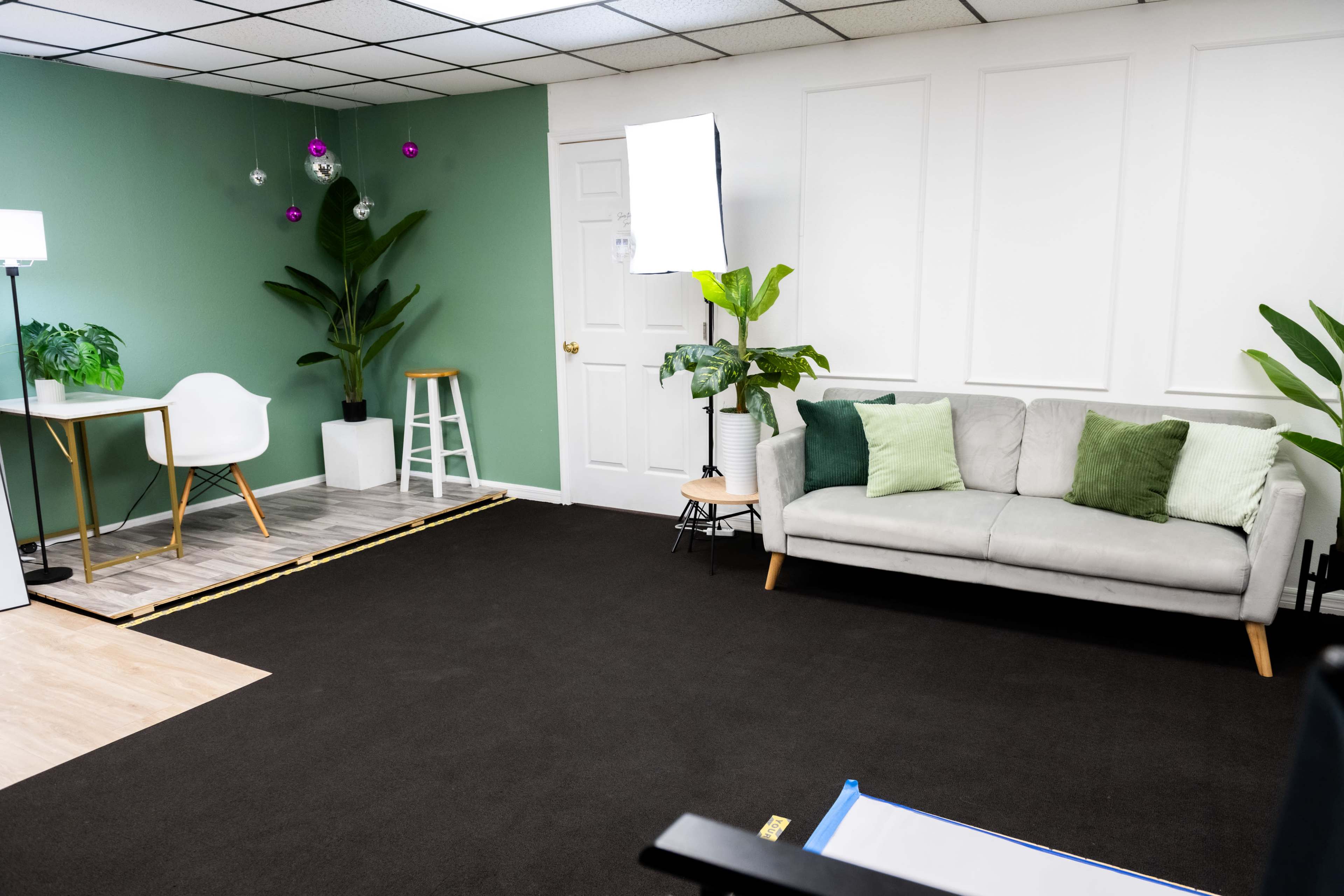 The image shows a modern room with a green accent wall, a gray sofa with decorative pillows, a small table with a white chair, and potted plants.