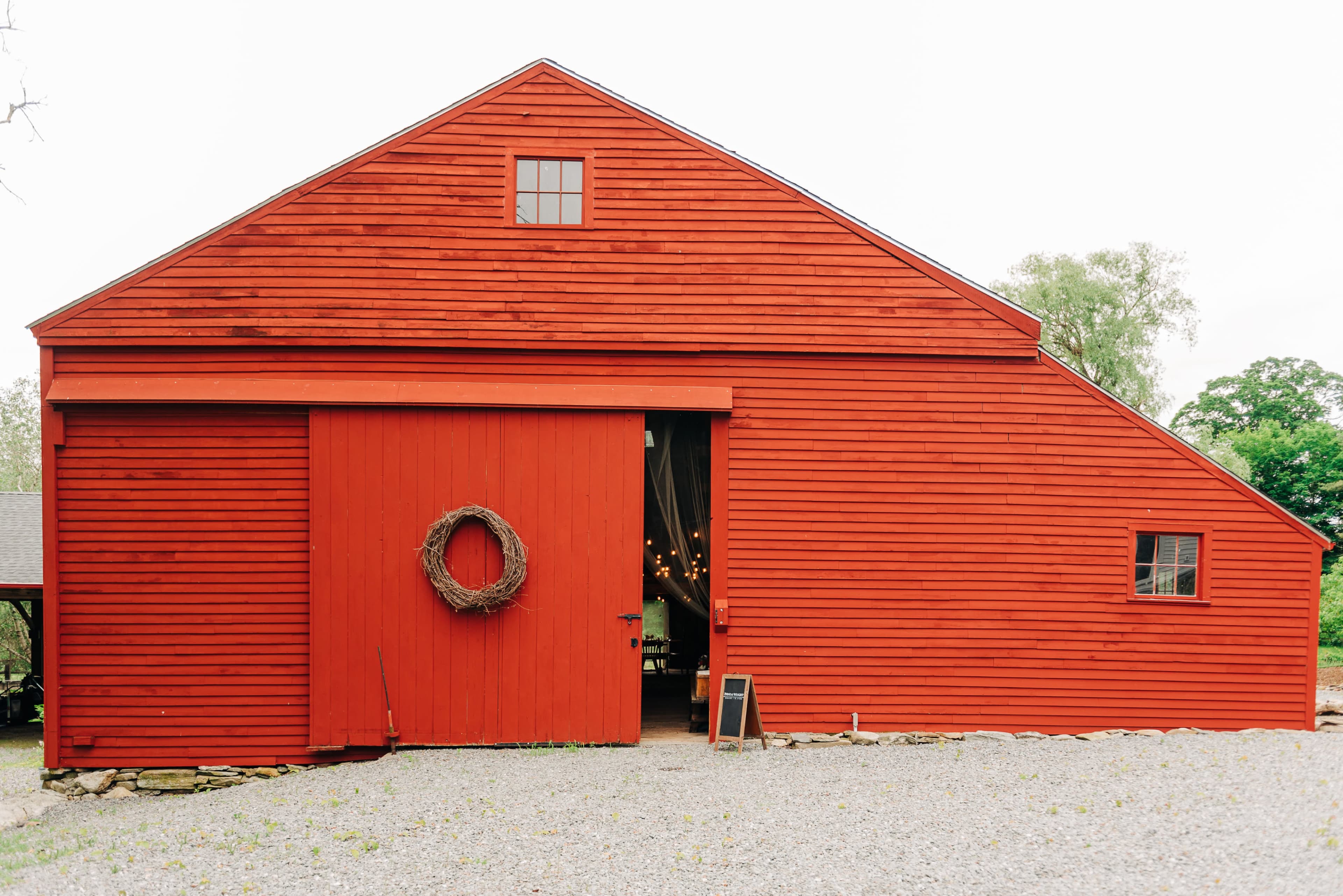 A large red barn with a door featuring a wreath is situated on a gravel path.