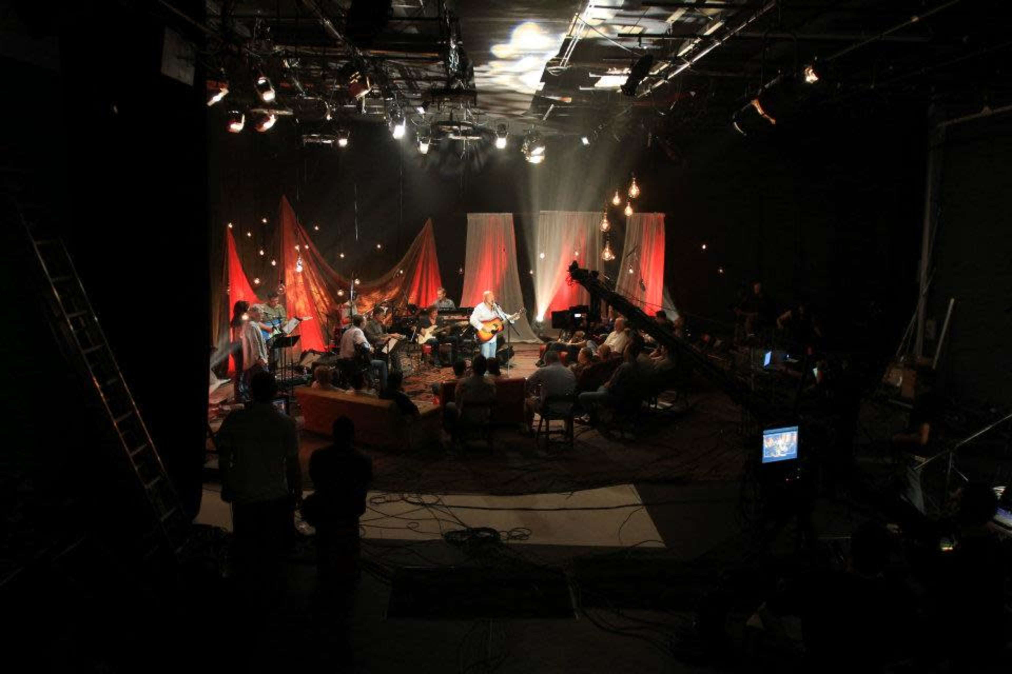 A live music performance is taking place on a dimly lit studio set with musicians and an audience arranged in a circular formation.