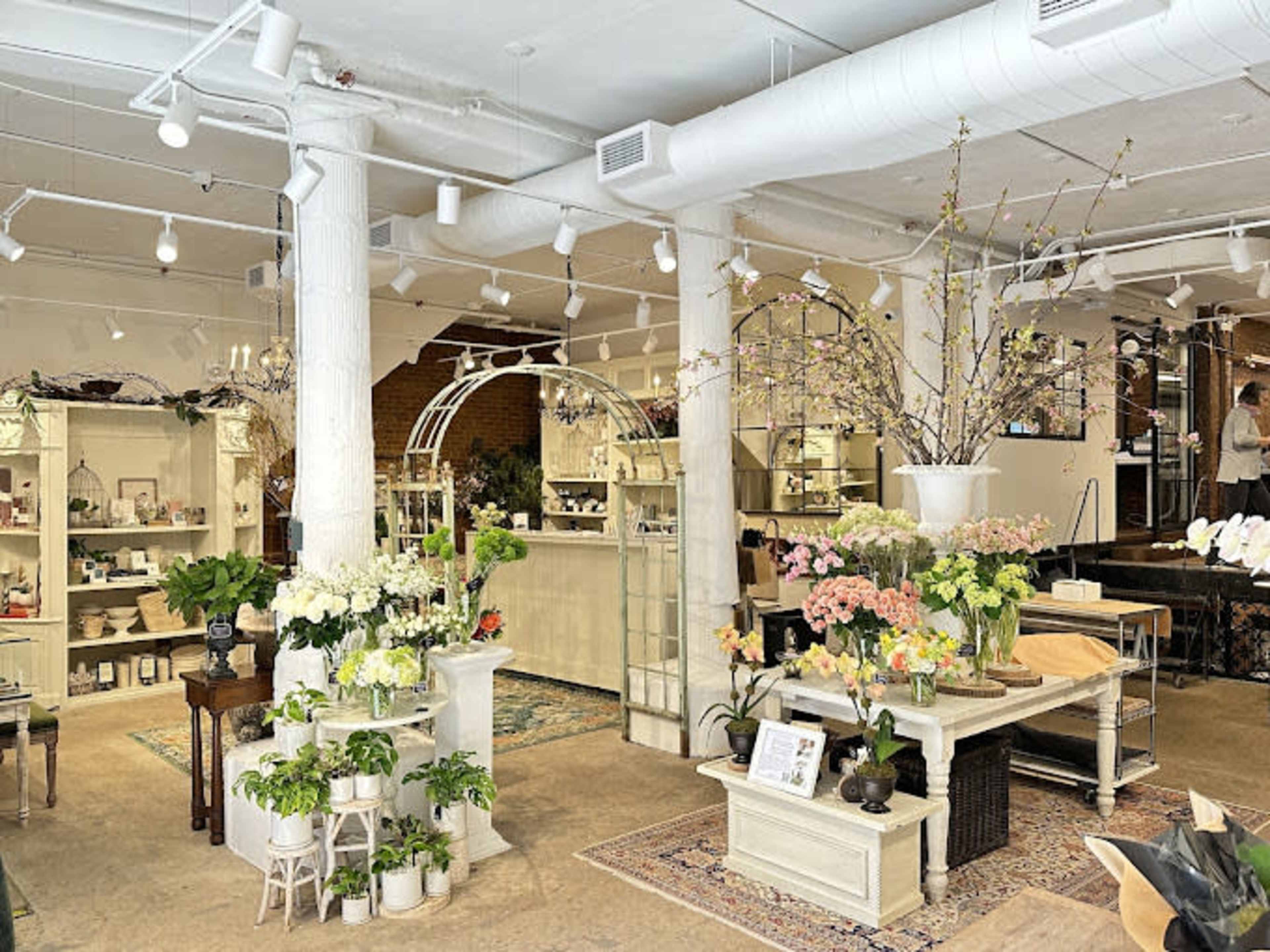 The image shows a well-organized flower shop featuring various floral arrangements displayed on tables, with shelves of plants and decorative items in the background.