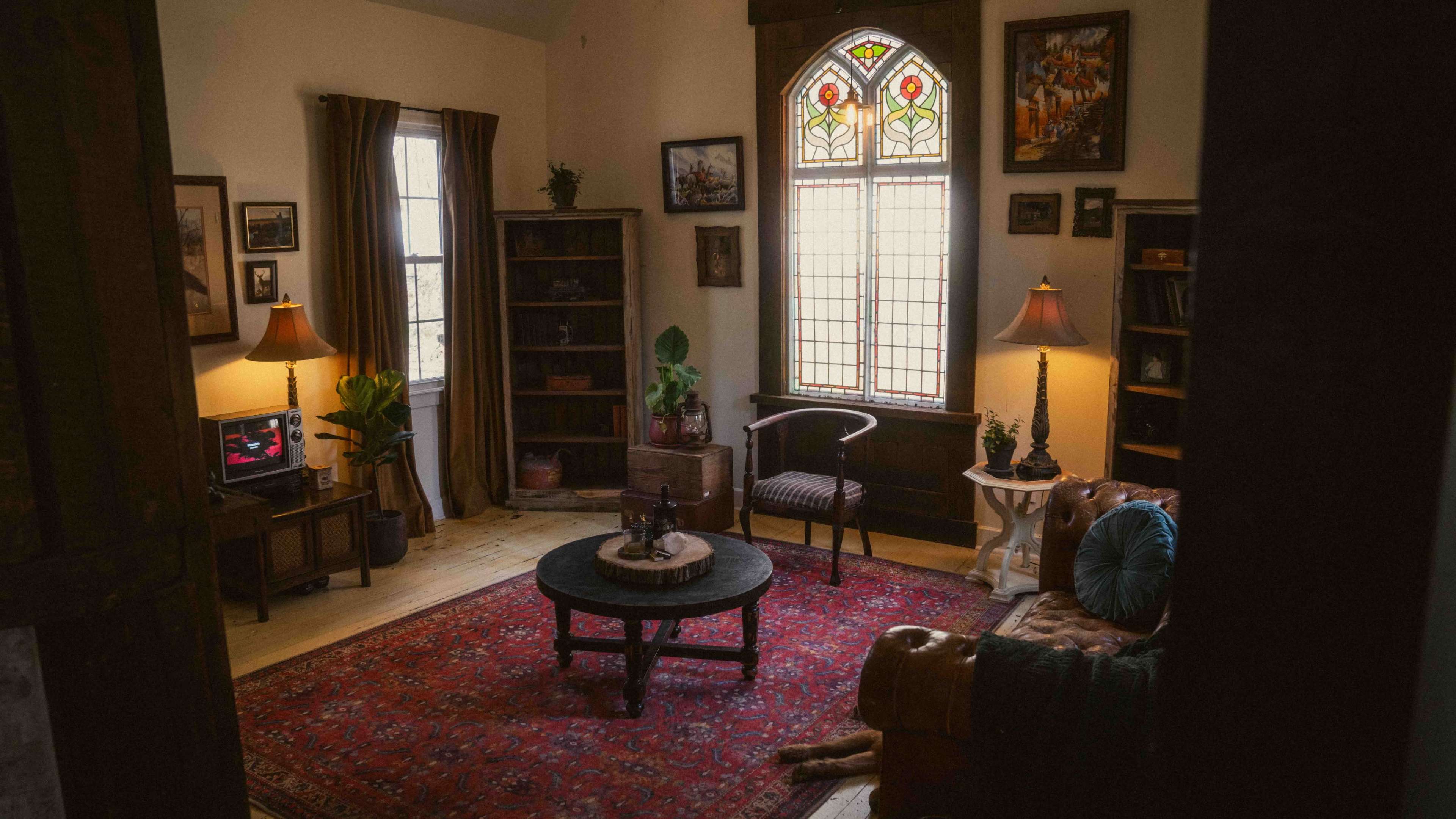 The image shows a cozy living room with a round table in the center, surrounded by a leather couch, bookshelves, and framed artwork on the walls.