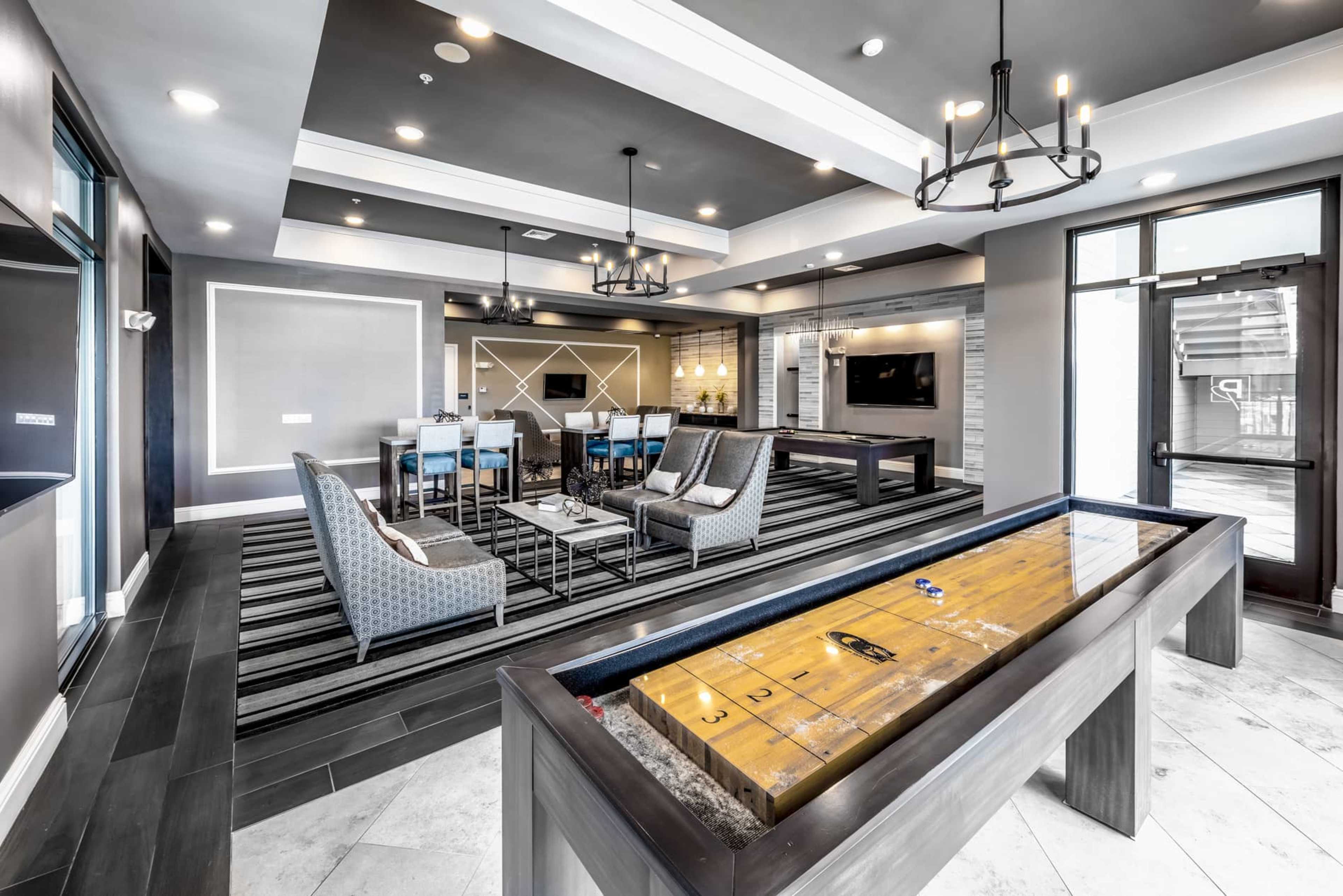 A modern lounge area featuring seating, a shuffleboard table, and a dining space with a television.