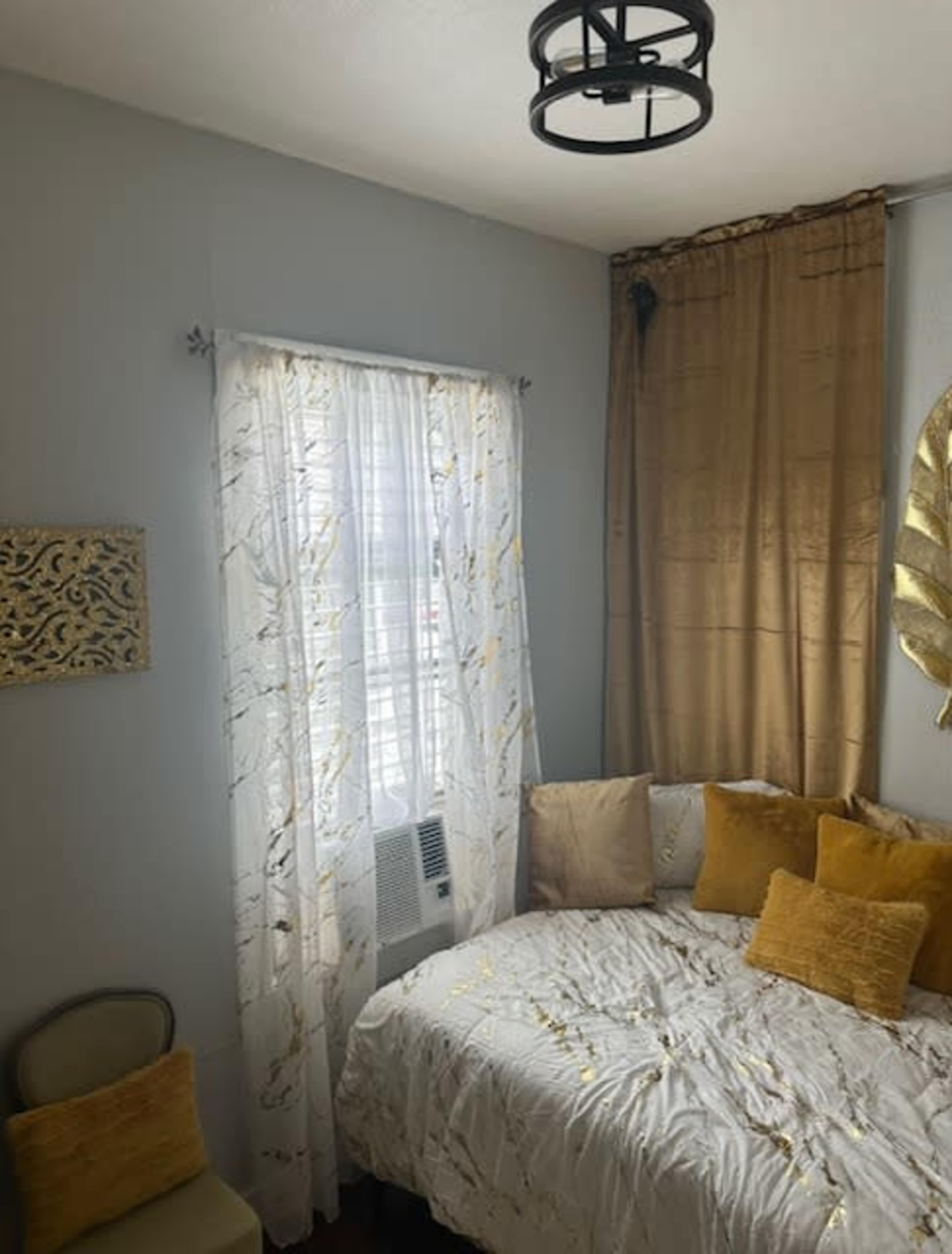 A small bedroom features a corner bed covered with a white and gold patterned comforter, surrounded by golden curtains and an air conditioning unit below a window.