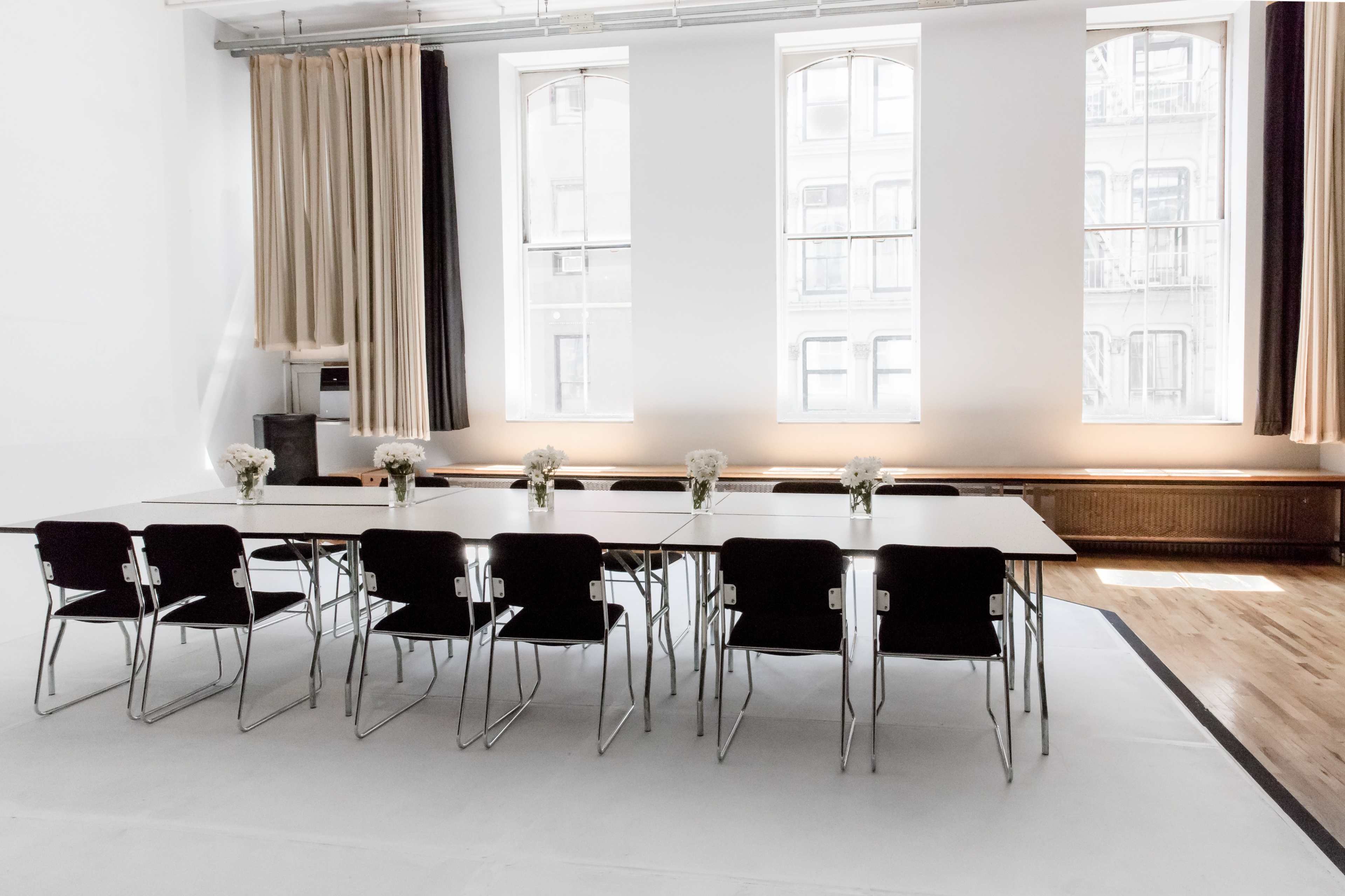 A long table with black chairs is set up in a bright, minimalist room with large windows and floral centerpieces.