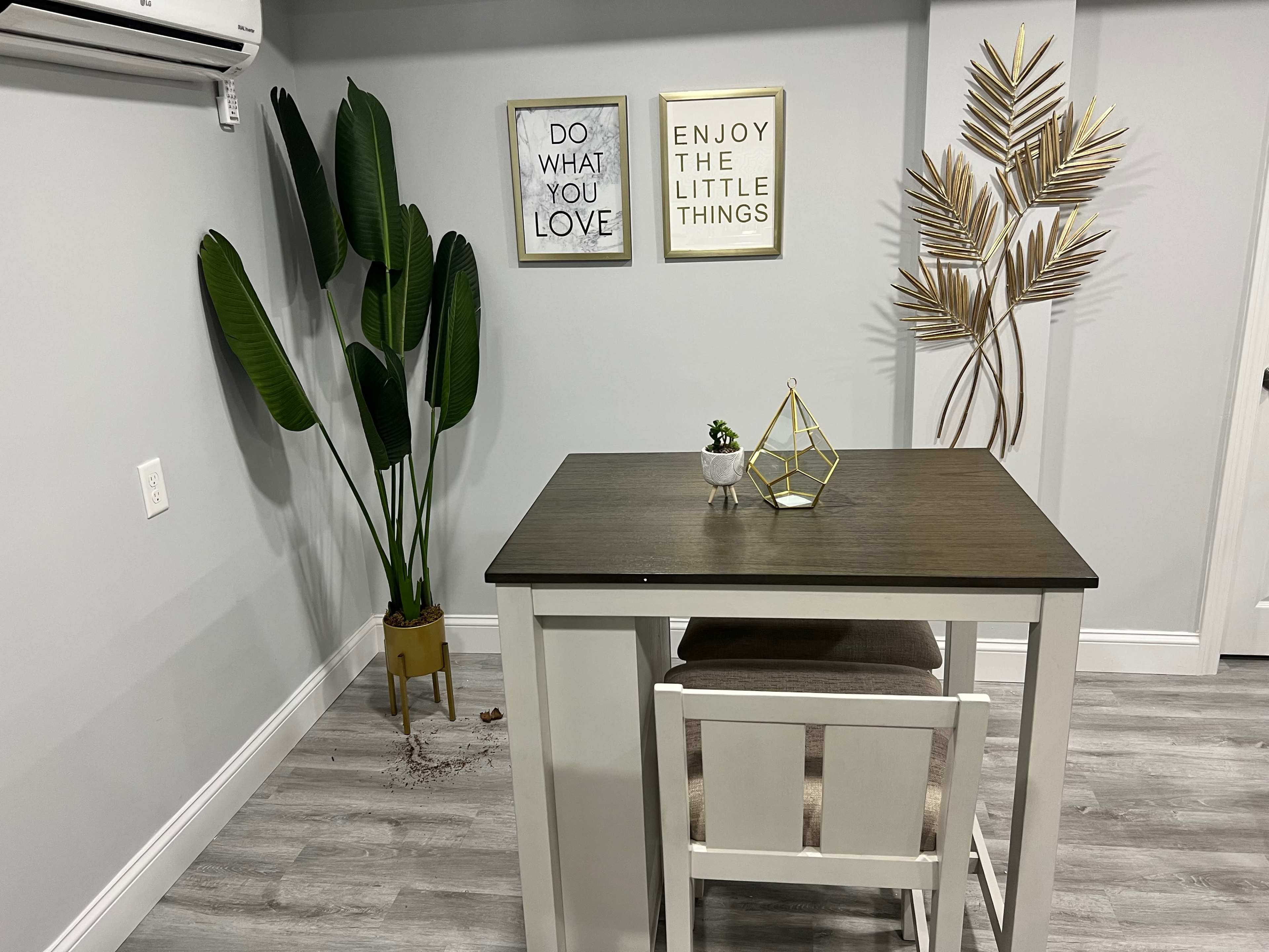 A small modern dining area with a table and chair, a decorative plant in a pot, and two framed motivational quotes on the wall.