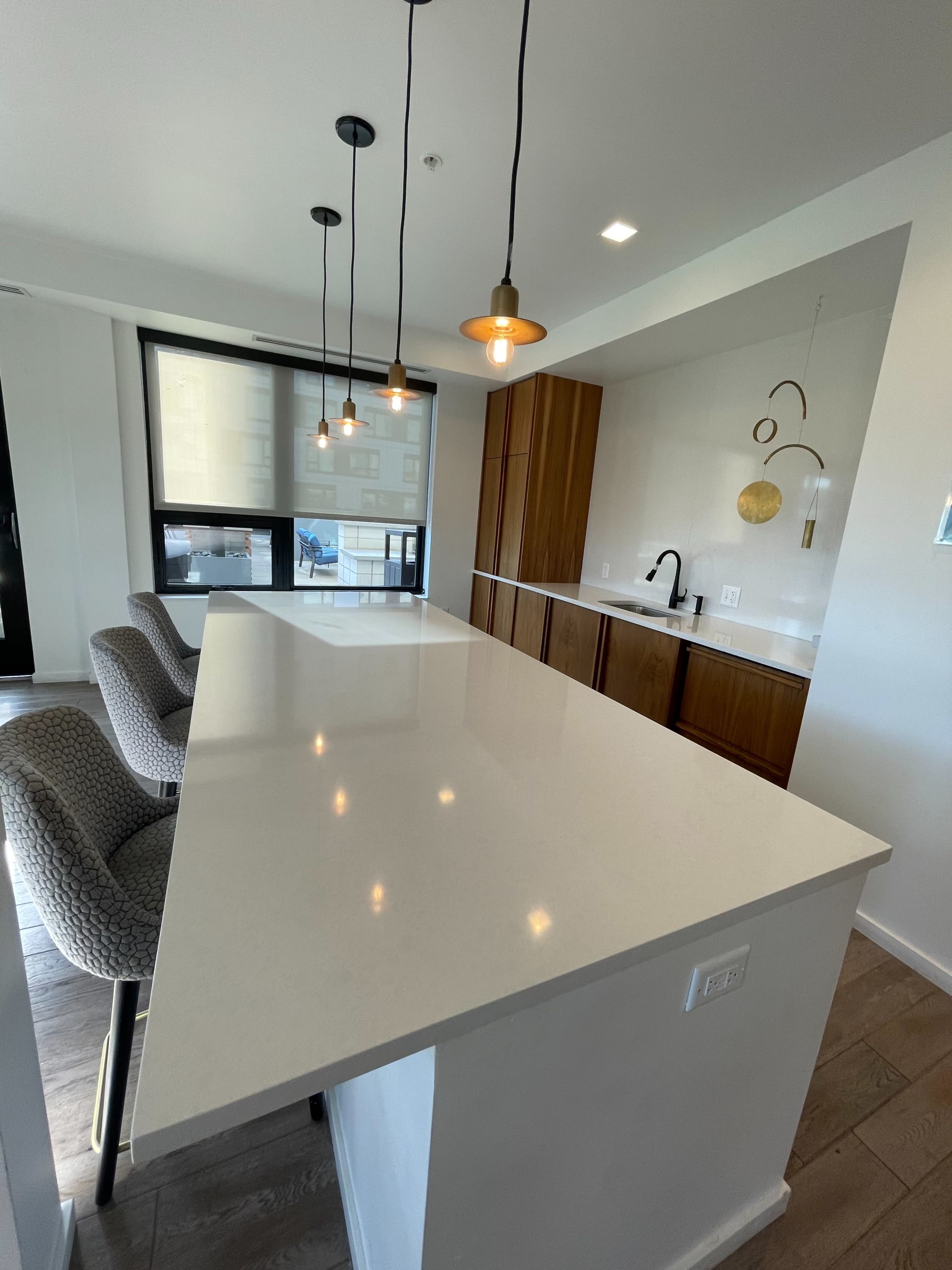 A modern kitchen features a large white countertop with two chairs and pendant lights, alongside wooden cabinetry and a window offering natural light.