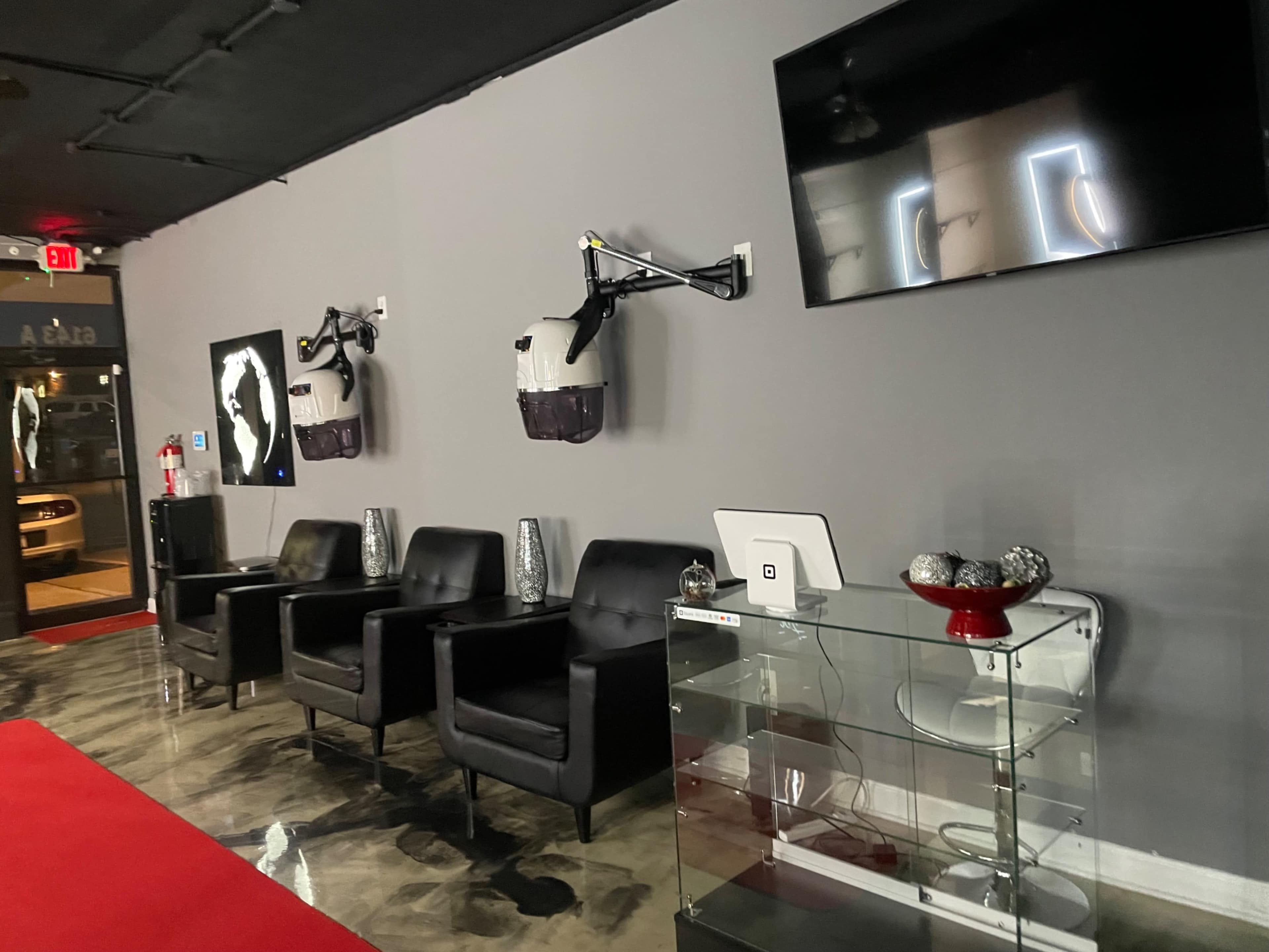 The image shows a modern waiting area with black chairs, sleek glass furniture, and a wall-mounted TV, featuring two mounted devices and artwork on the walls.