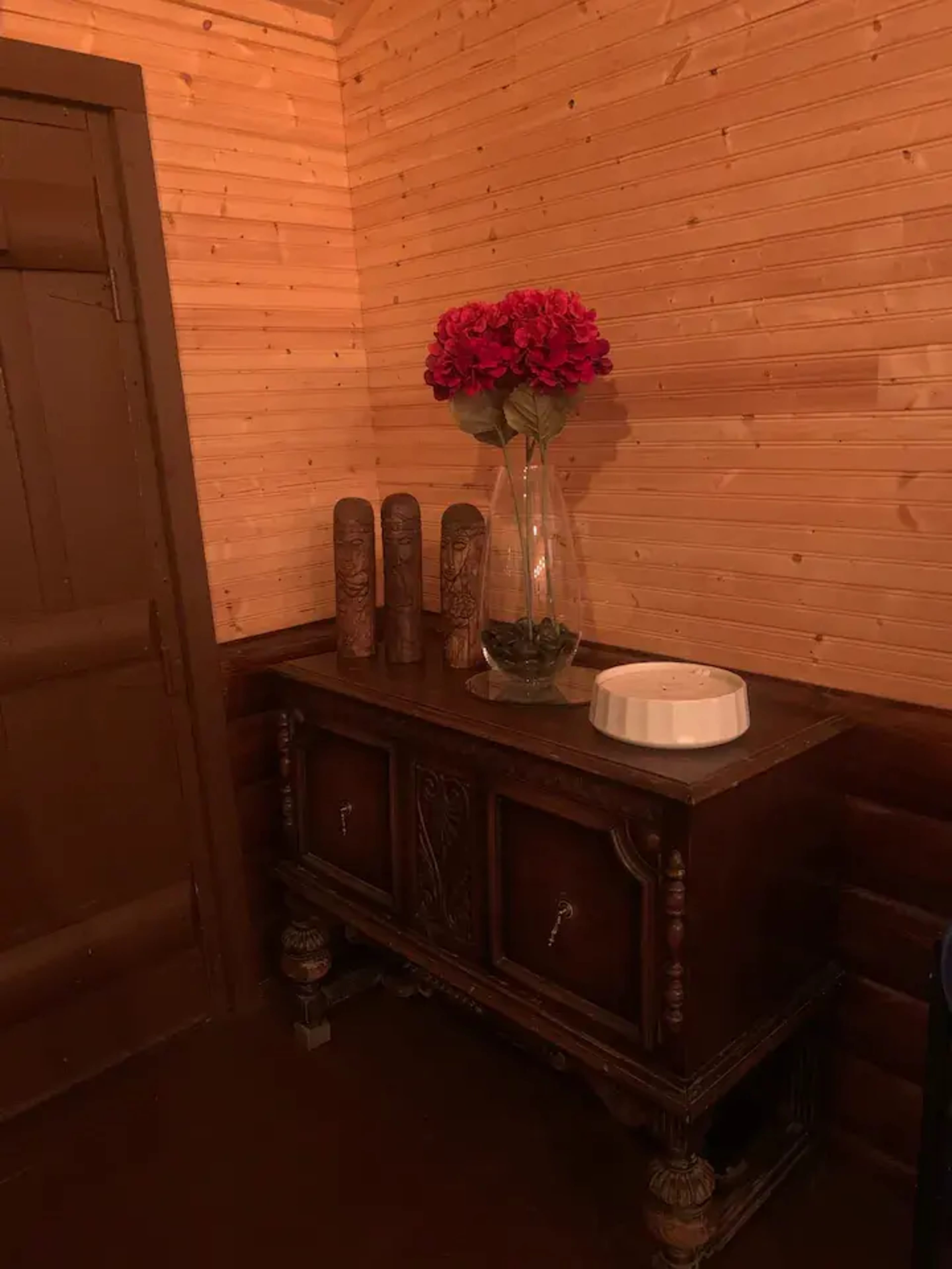 A wooden sideboard decorated with a glass vase of flowers and three carved pillars stands against a wooden wall in a dimly lit room.