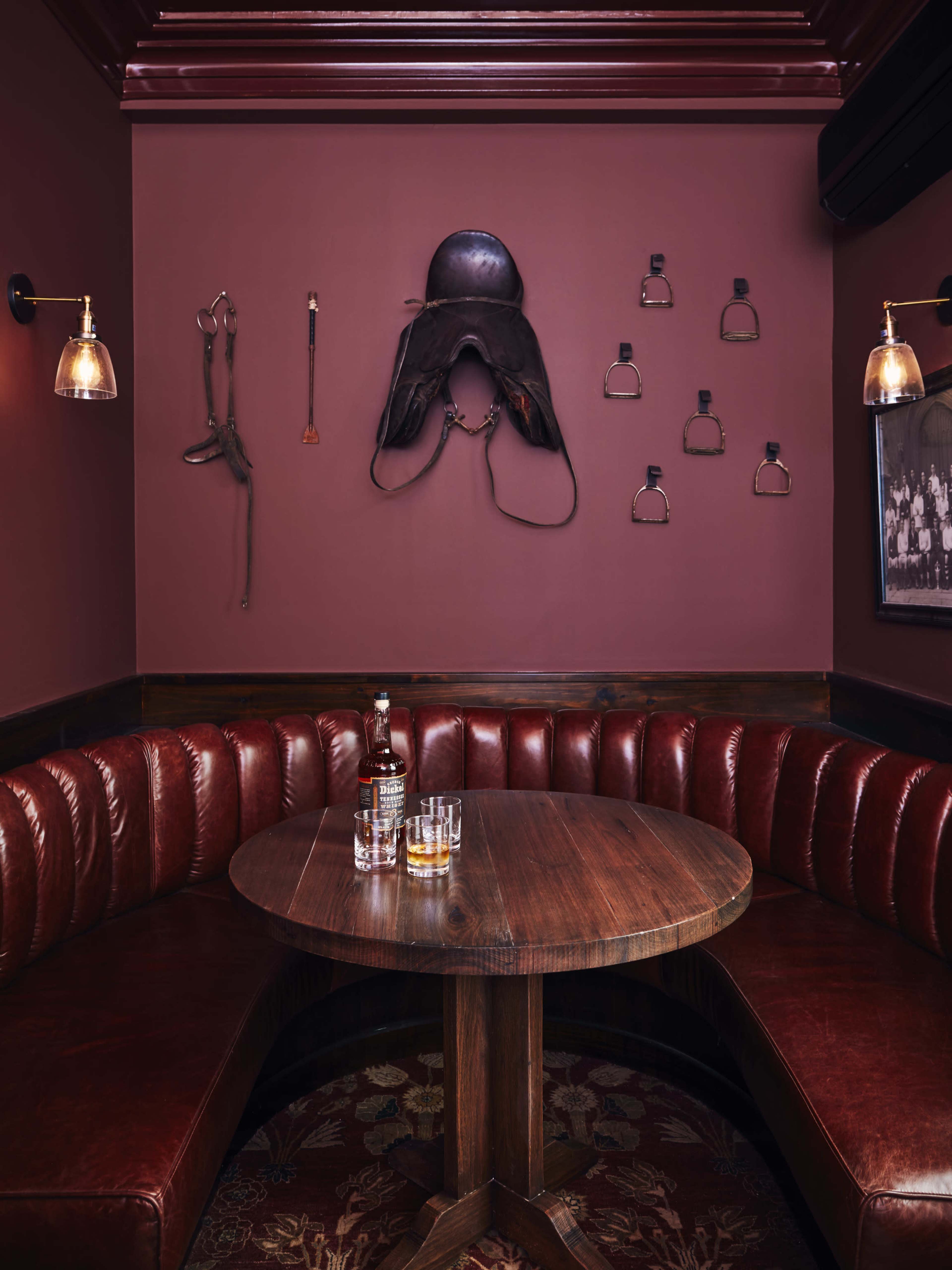 A cozy booth in a dimly lit restaurant features a round wooden table, surrounded by padded red leather seating, with equestrian-themed decor on the wall.