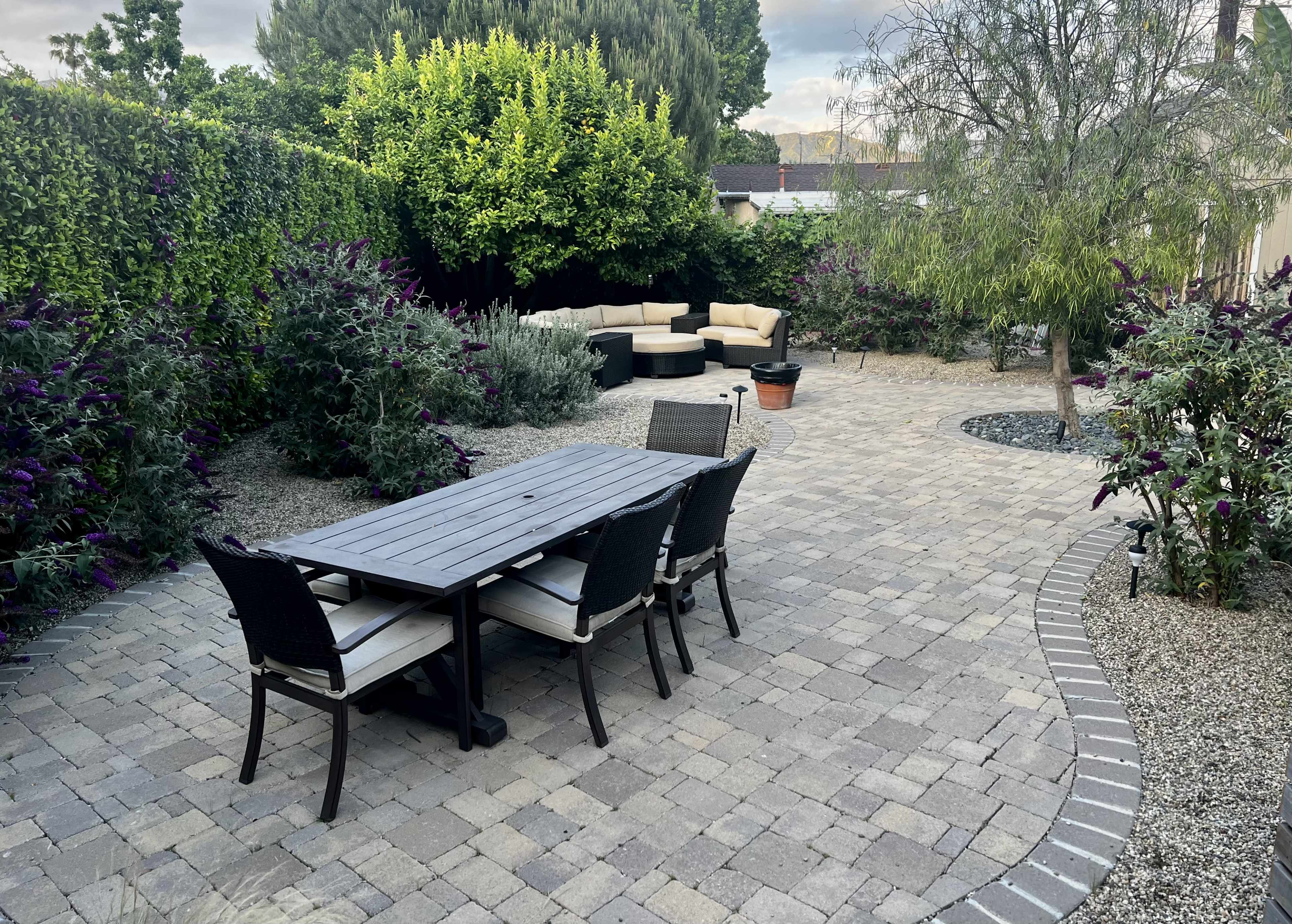 A paved patio area features a rectangular table with chairs, surrounded by garden plants and a sectional seating arrangement.