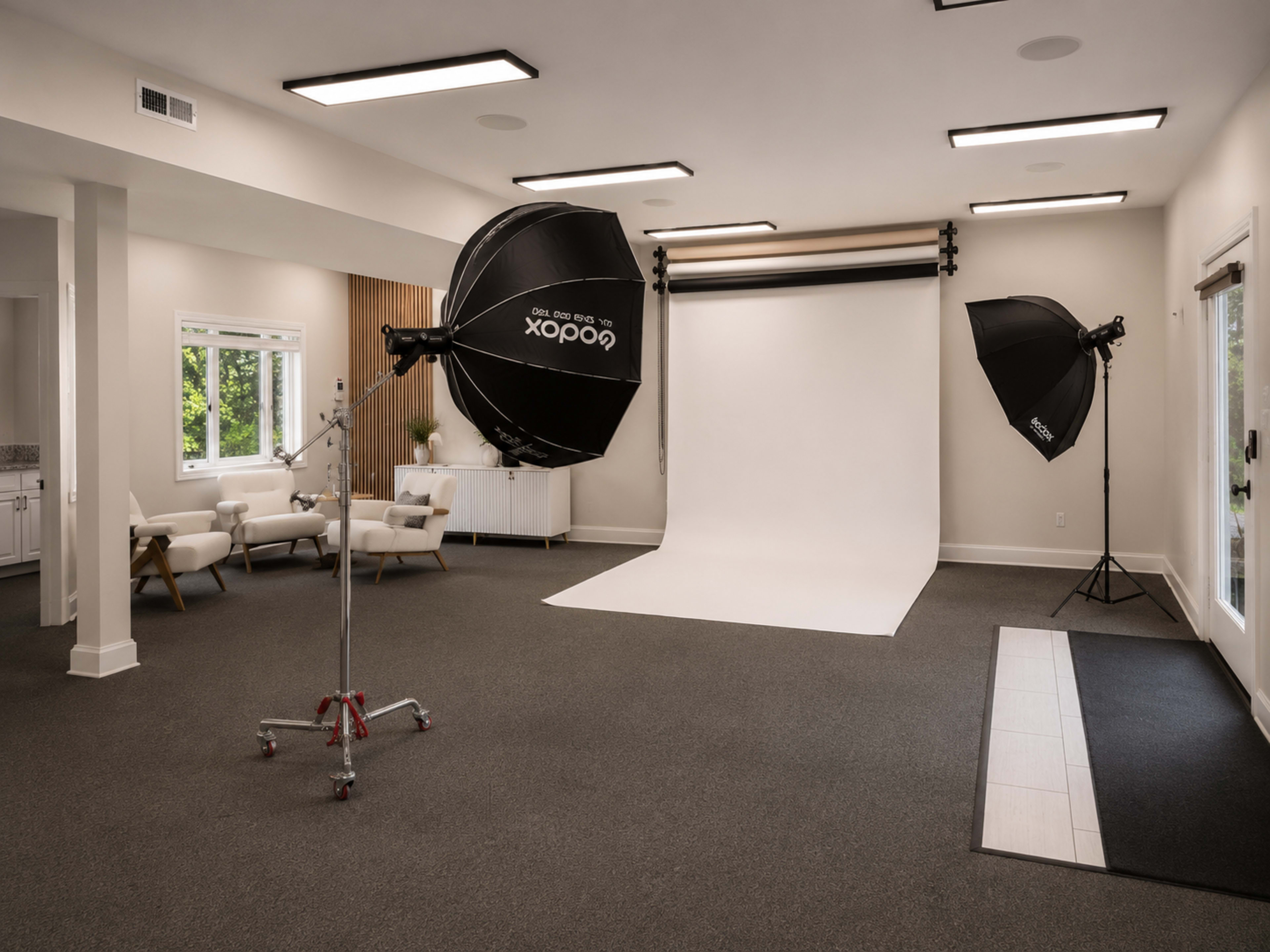 The image shows a well-lit photography studio featuring a plain white backdrop, two large softbox lights, and modern seating in a corner.
