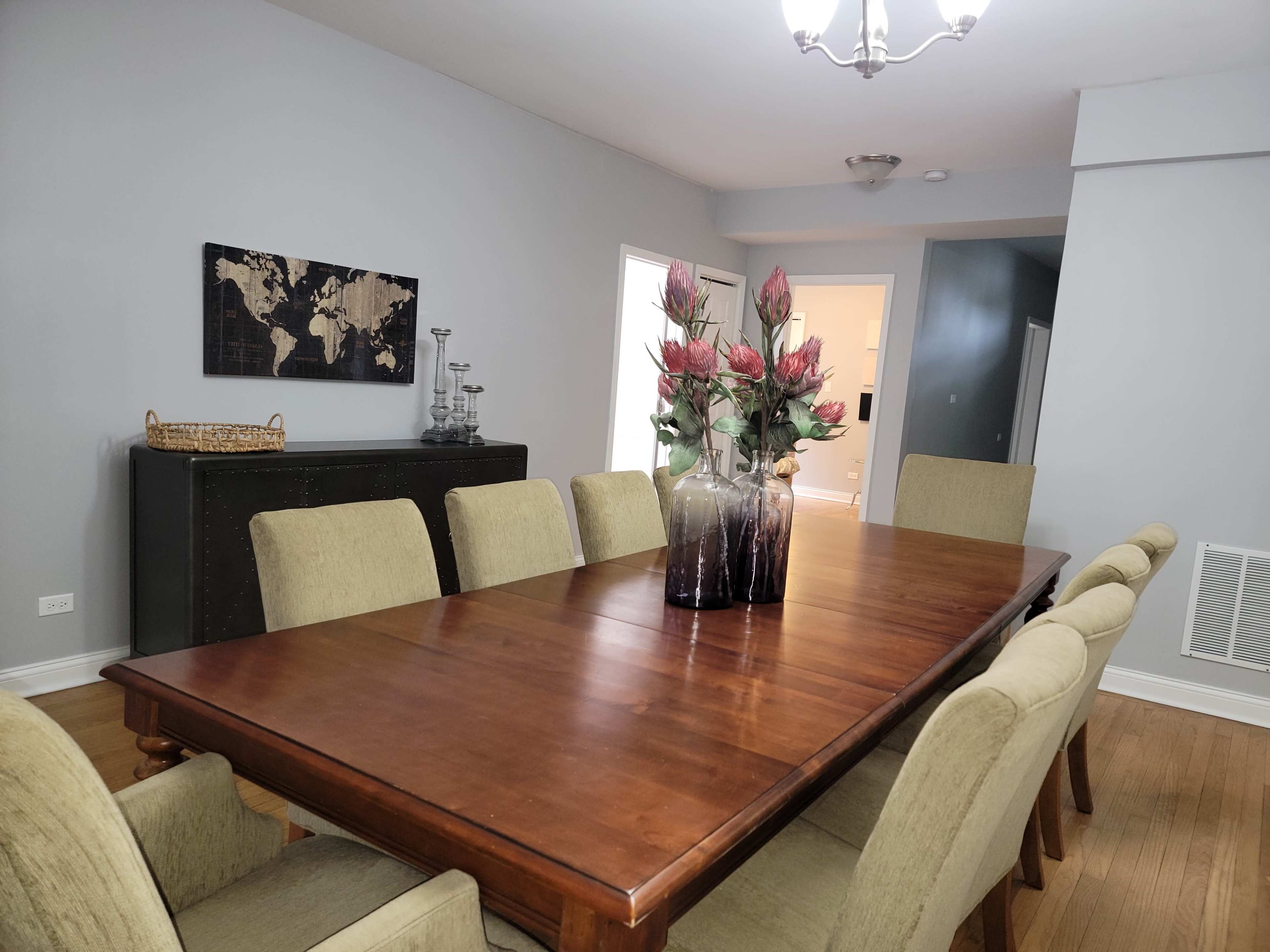 The image shows a spacious dining room featuring a large wooden table surrounded by upholstered chairs, with a decorative vase of flowers as the centerpiece.