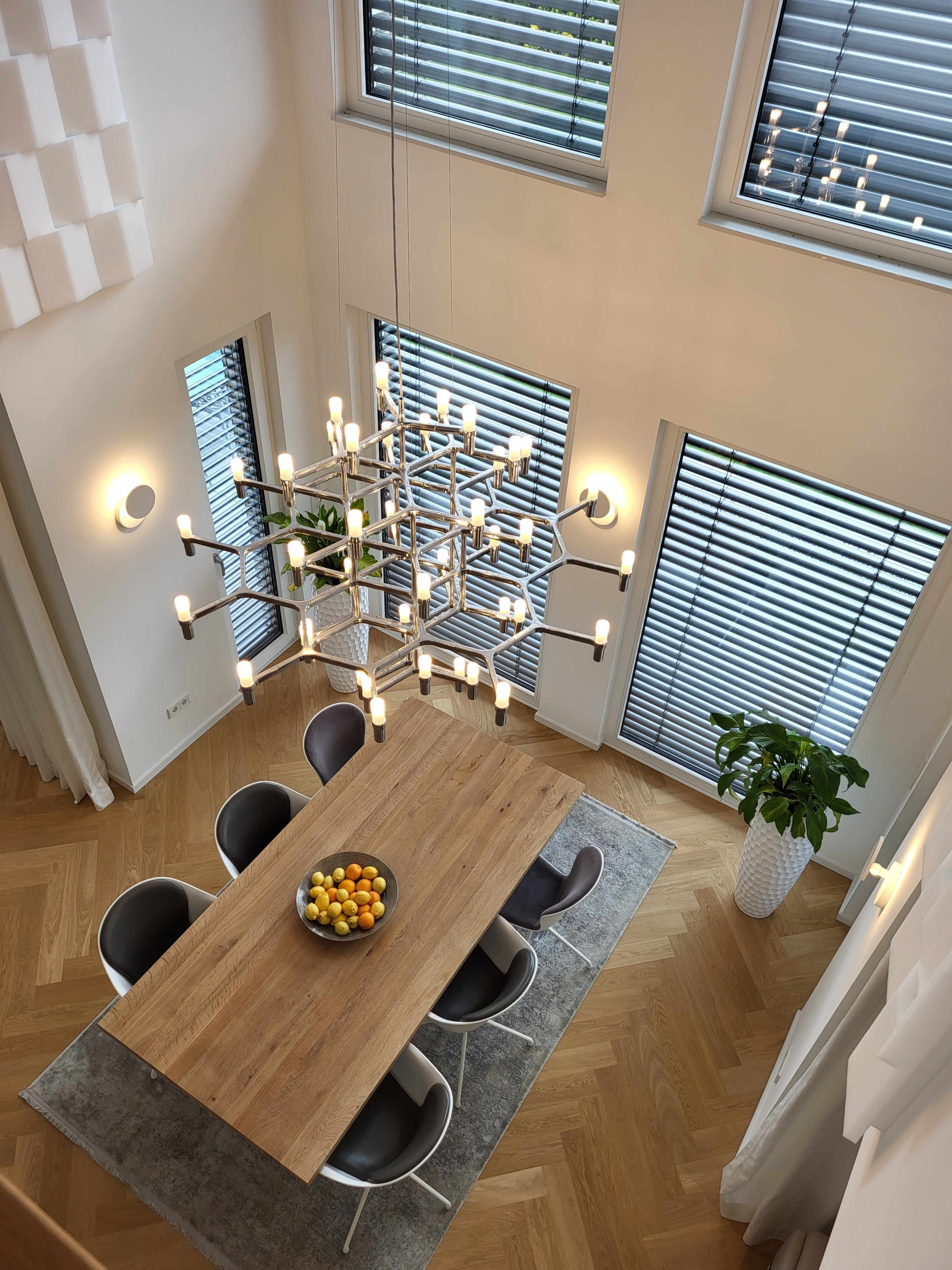 A modern dining area features a large wooden table surrounded by six chairs, illuminated by a geometric chandelier, with large windows covered by blinds and a decorative plant in the corner.