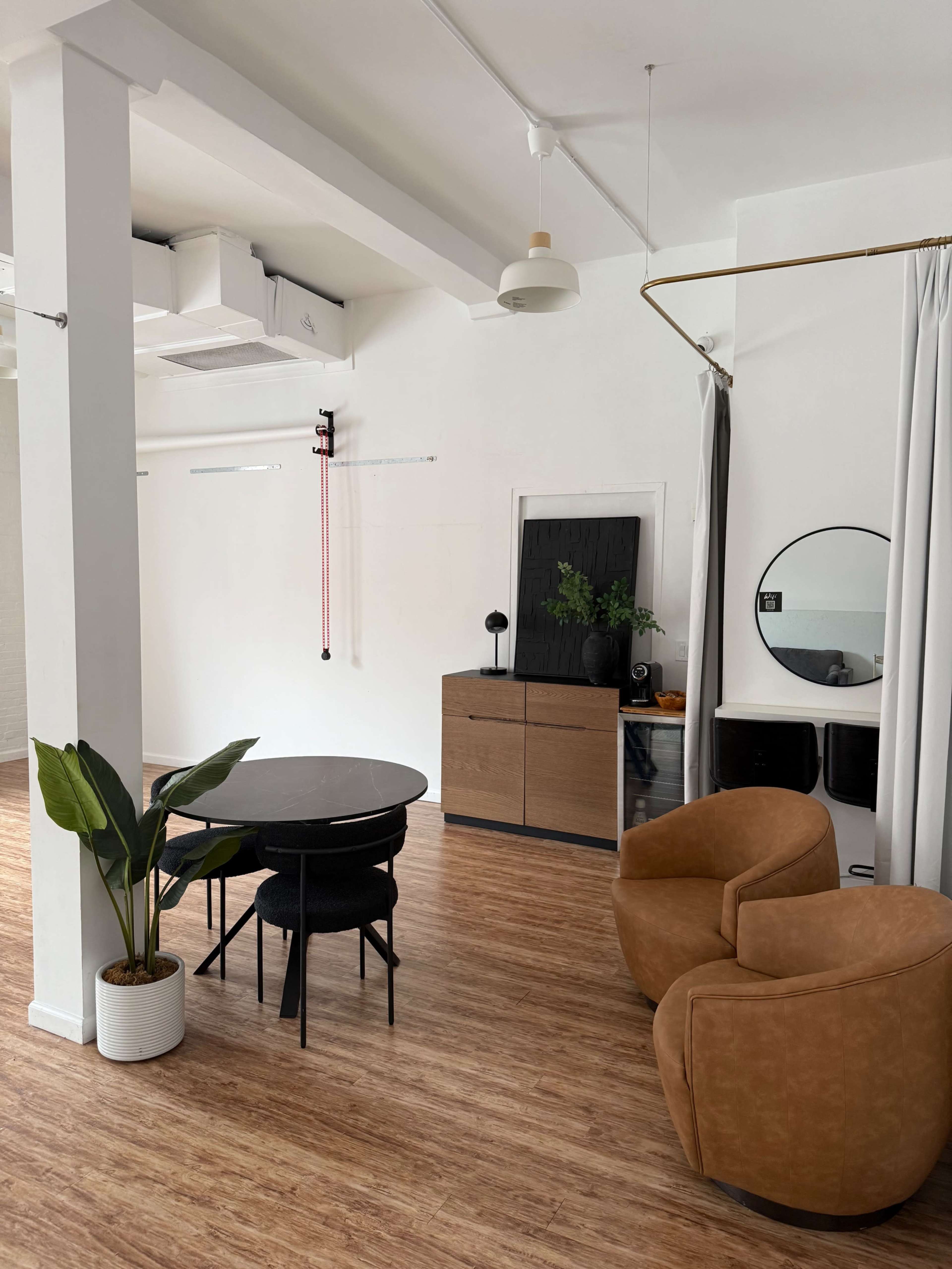 The image shows a modern interior space featuring a black circular dining table with four chairs, a light wooden floor, a brown accent chair, and a wall-mounted mirror.