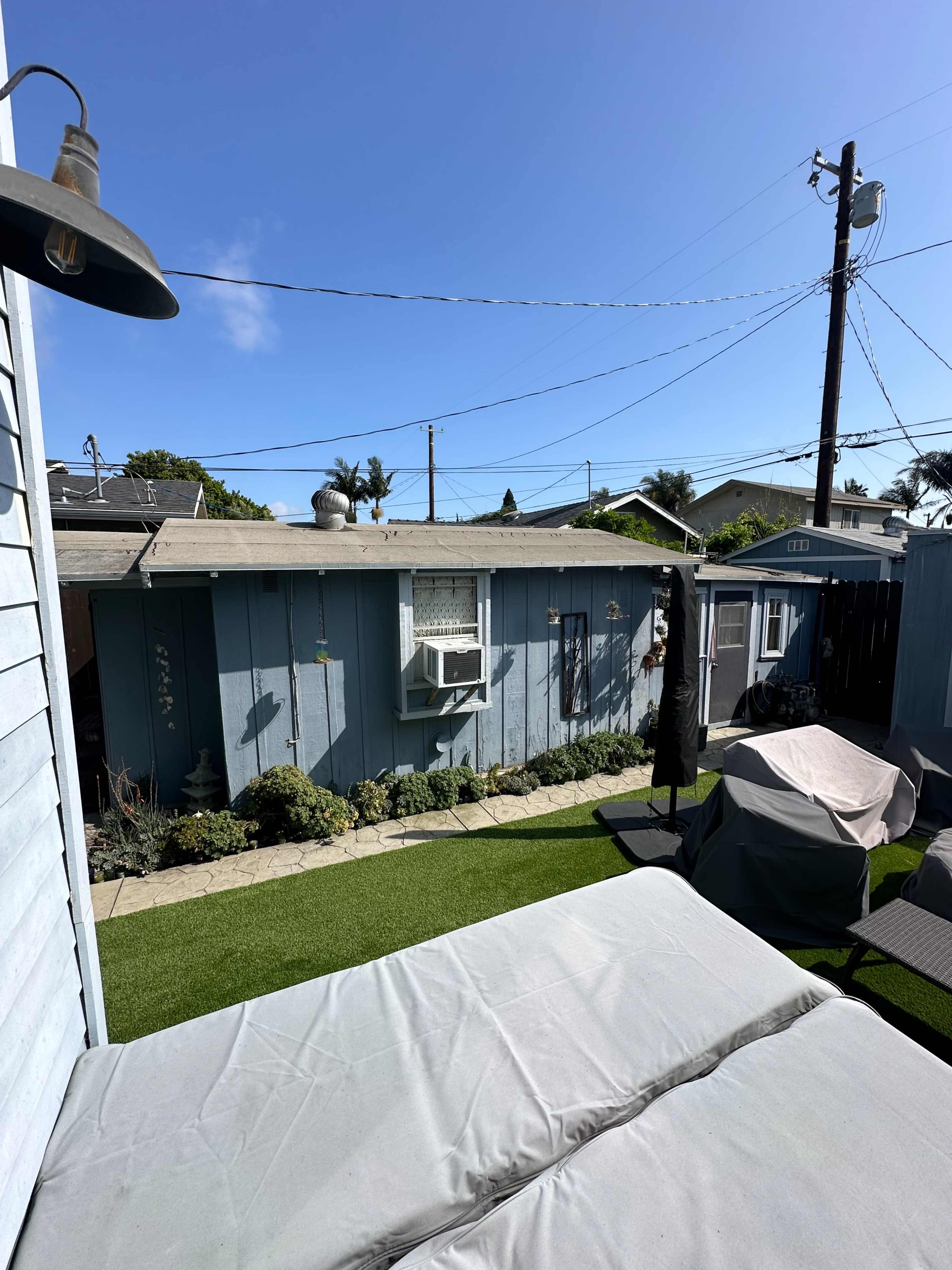 The image shows a backyard with a blue shed, air conditioning unit, and patio furniture on artificial grass under a clear blue sky.