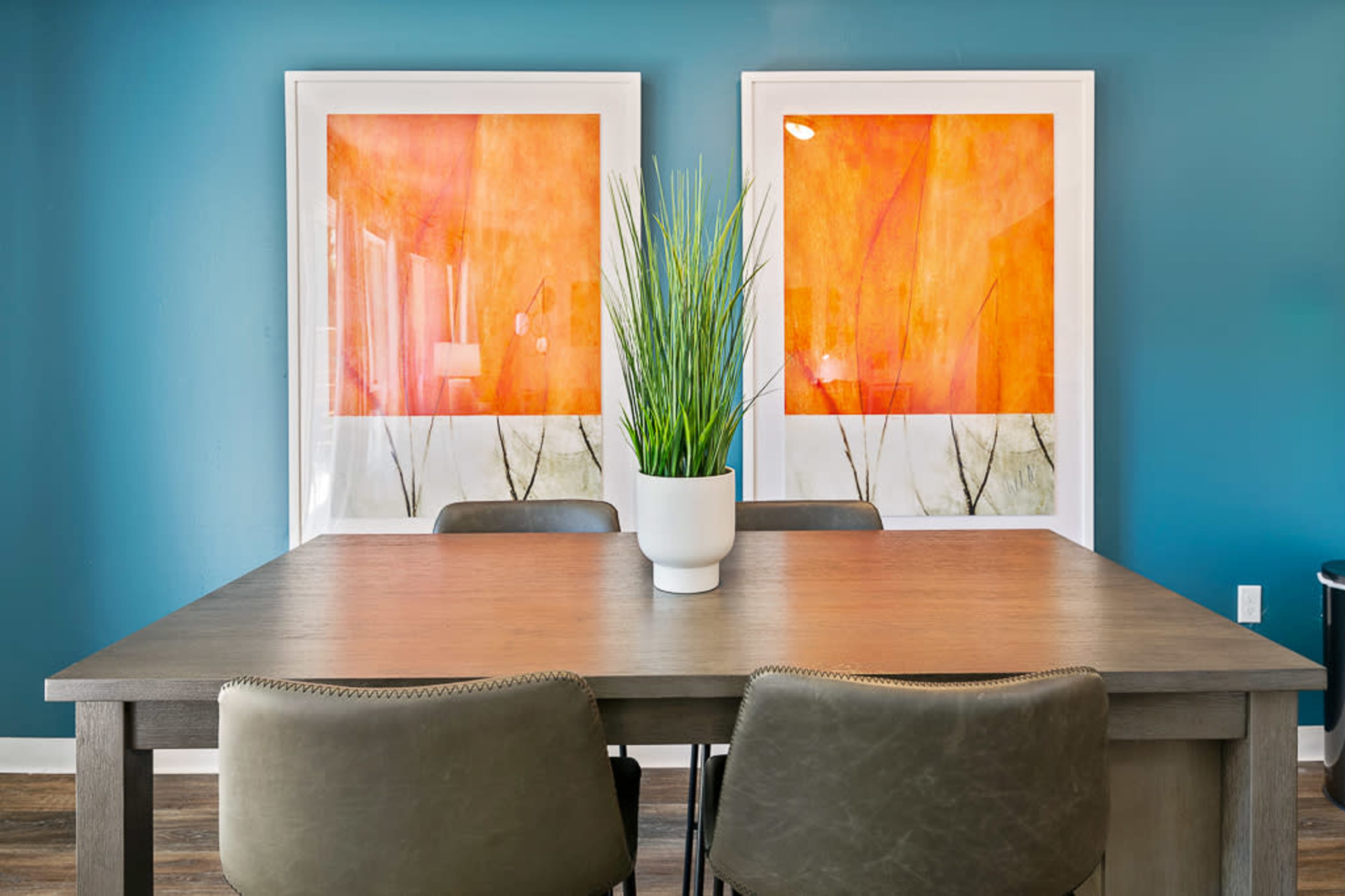 A modern dining table with three chairs is positioned in front of two colorful framed artworks against a blue wall, with a potted plant centerpiece.