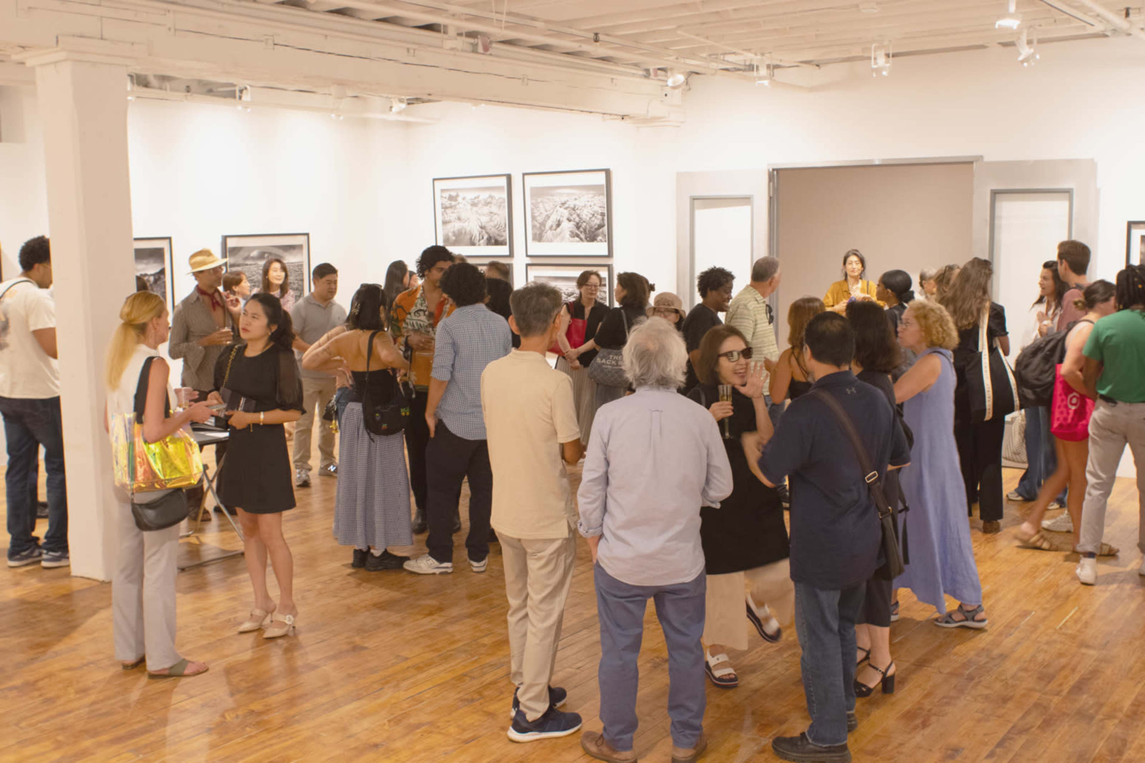 A crowd of people mingles at an art gallery opening, surrounded by large black-and-white photographs on the walls.